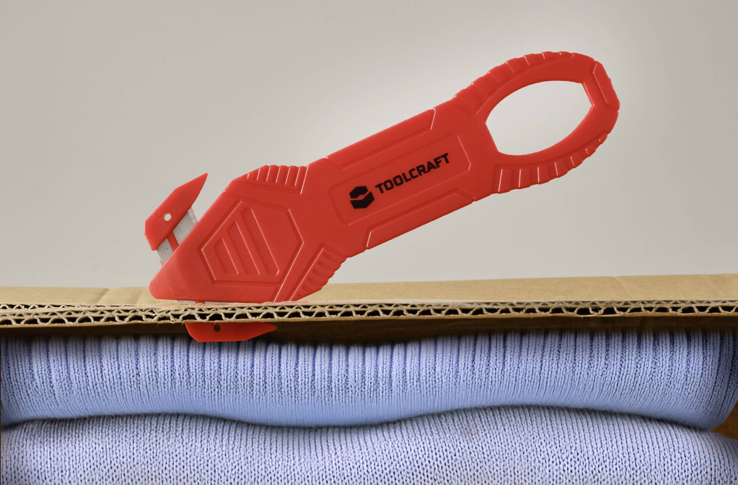 Ein roter Teppichmesser mit der Aufschrift "TOOLCRAFT" schneidet in einen Stapel Pappe, der über zusammengelegten Kleidungsstücken positioniert ist.