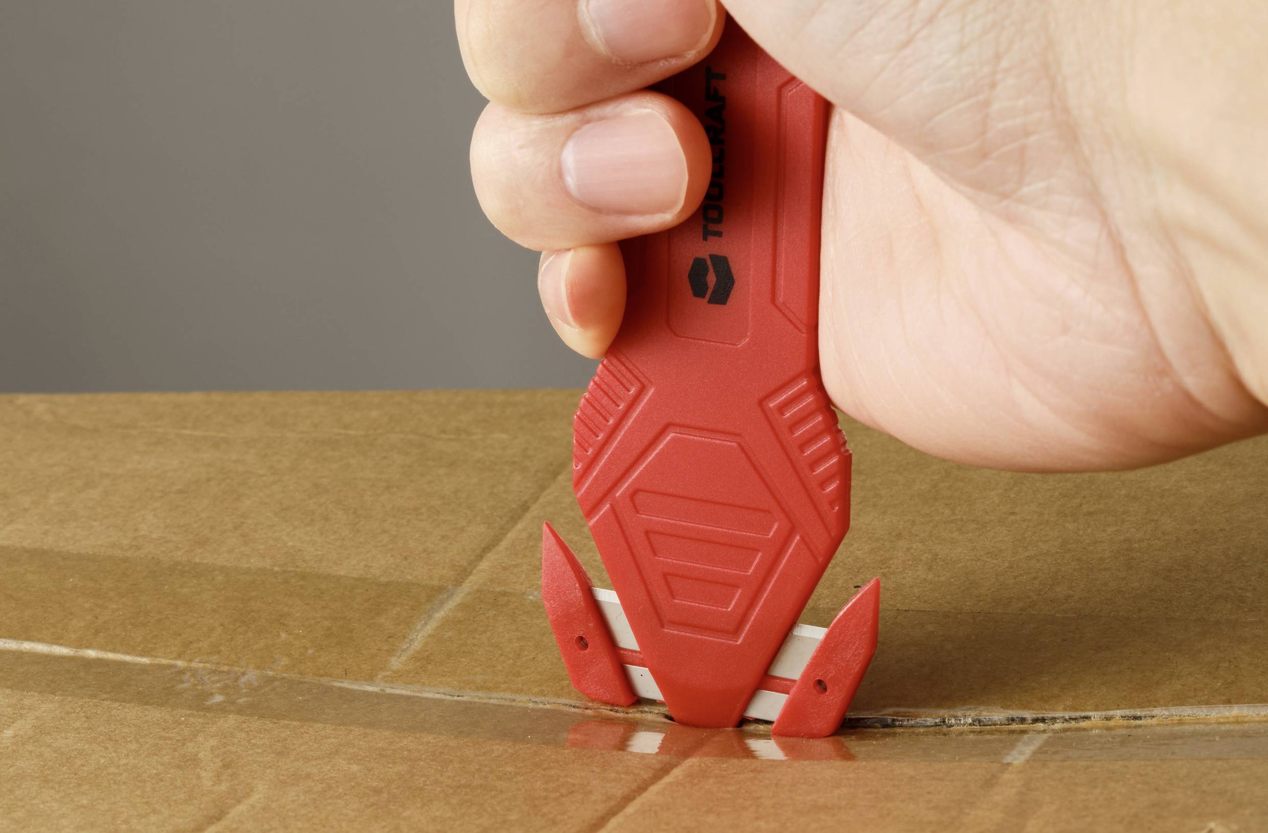 Eine Hand schneidet mit einem roten Sicherheits-Cuttermesser durch Karton und demonstriert das sichere Öffnen eines versiegelten Pakets.