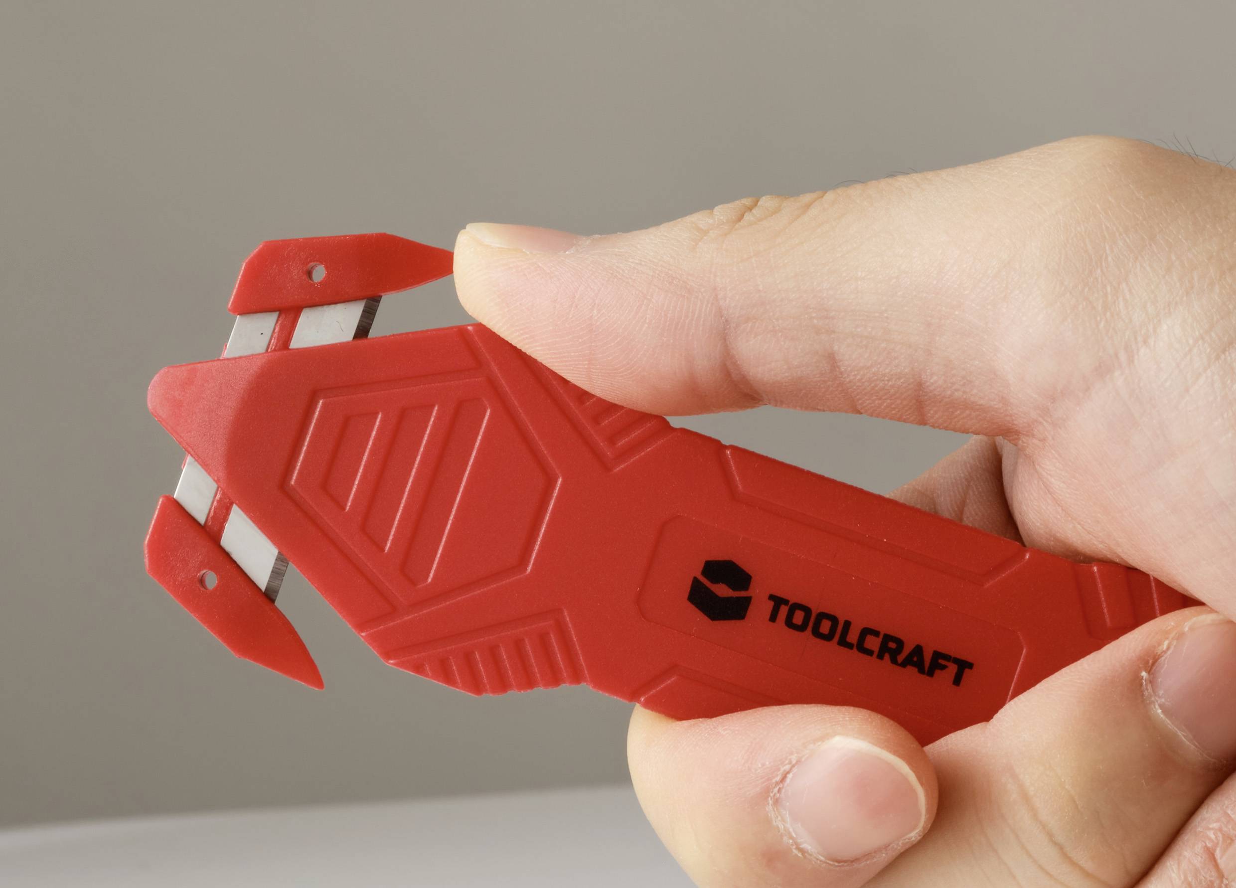 Eine Hand, die ein rotes Werkzeug mit der Aufschrift "TOOLCRAFT" hält, das am Ende eine Klinge aufweist und vermutlich für Schneid- oder Abstreifaufgaben konzipiert ist.