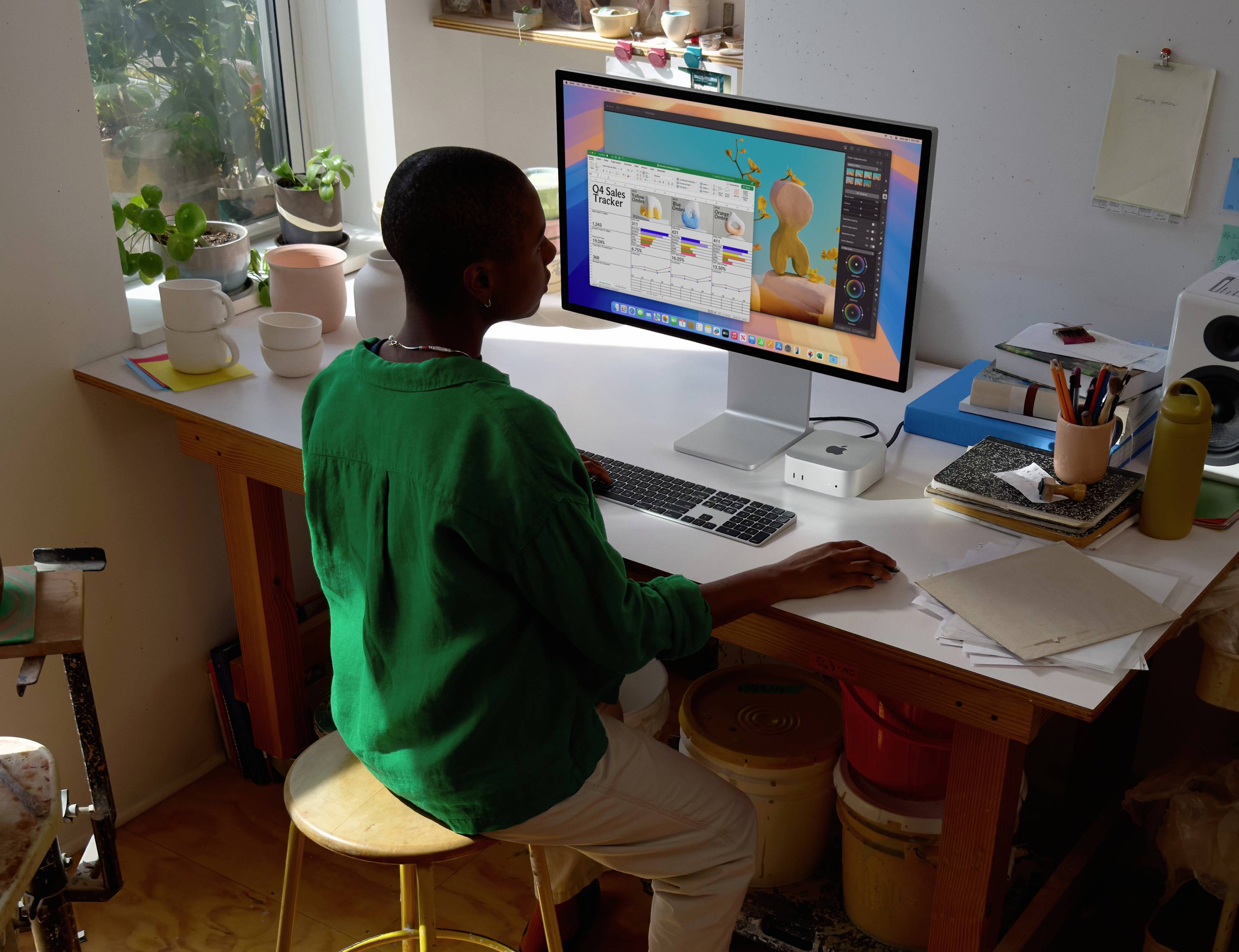 Eine Person in einem grünen Shirt sitzt an einem Schreibtisch und arbeitet an einem Computer, umgeben von Künstlerbedarf und Pflanzen, während auf dem Bildschirm eine Designsoftware geöffnet ist.