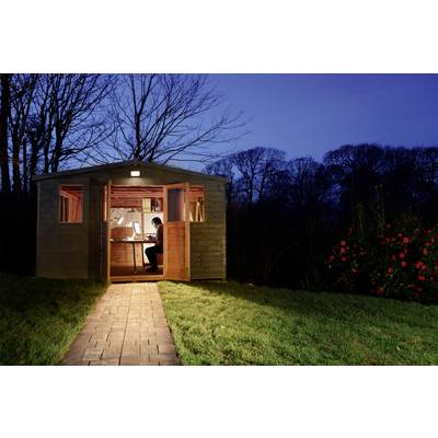 Eine Person arbeitet nachts in einem beleuchteten Schuppen im Garten. Die Tür ist offen, und der Schuppen ist von Bäumen umgeben.
