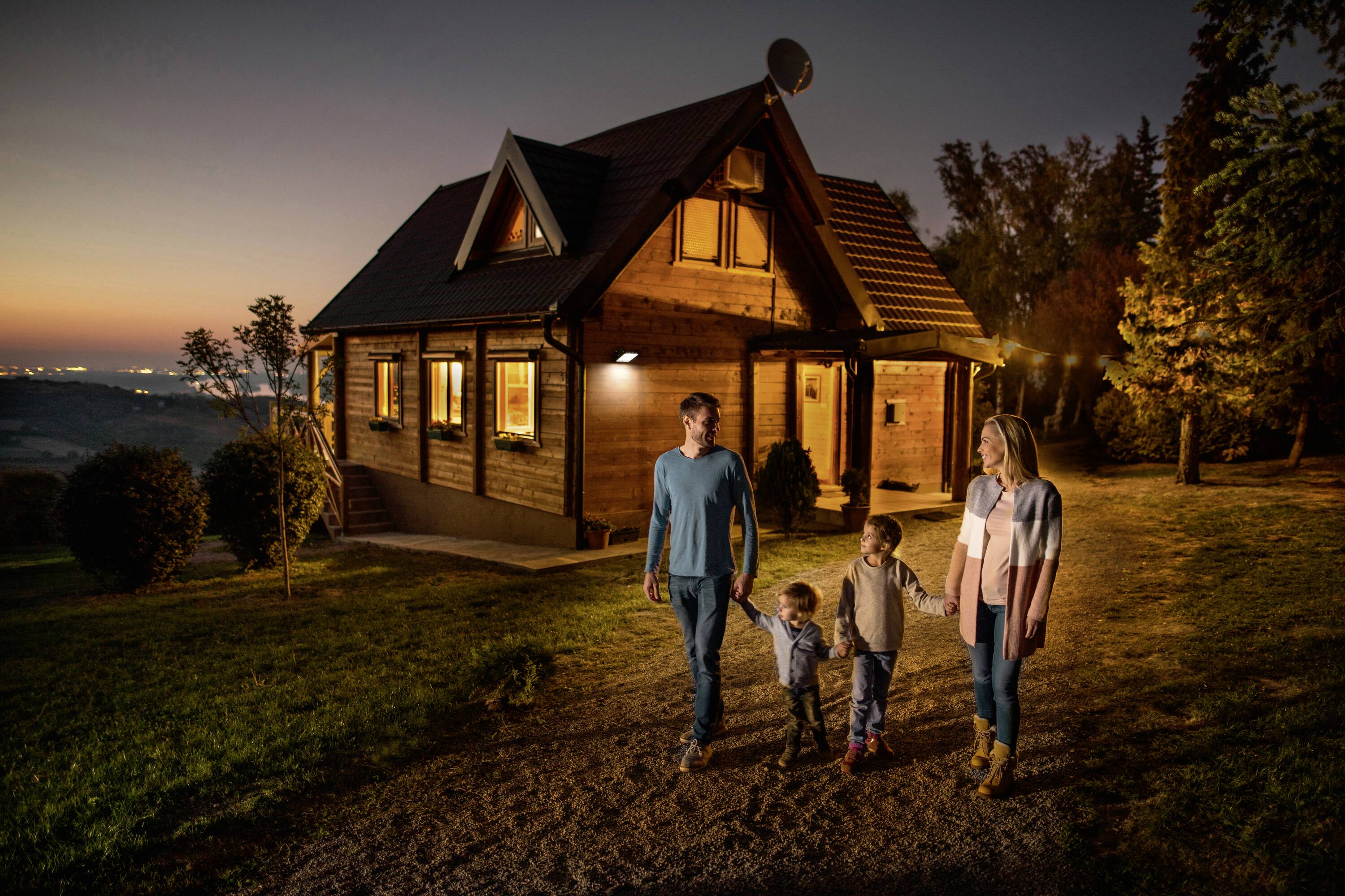 Eine Familie mit zwei Kindern spaziert bei Sonnenuntergang auf einem Weg vor einem Holzhaus. Die Szene strahlt Ruhe und Harmonie aus.