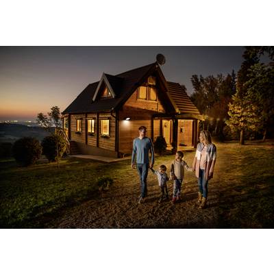 Eine Familie mit zwei Kindern spaziert bei Sonnenuntergang auf einem Weg vor einem Holzhaus. Die Szene strahlt Ruhe und Harmonie aus.