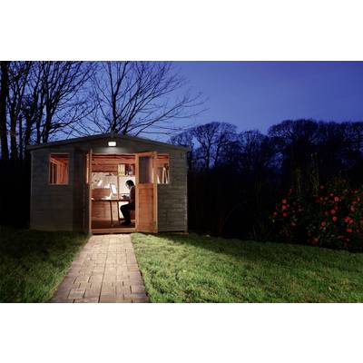 Eine Person arbeitet abends an einem Schreibtisch in einem beleuchteten Gartenschuppen. Draußen ist es dunkel, der Weg ist beleuchtet.