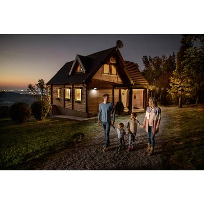 Eine Familie spaziert bei Sonnenuntergang auf einem Weg zu einem Holzhaus. Die Umgebung ist grün und bewaldet.