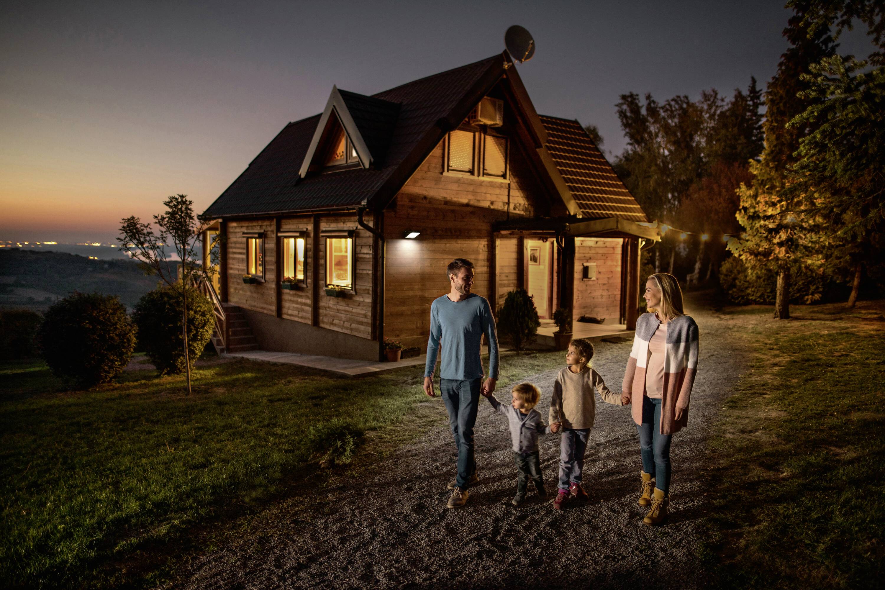 Eine Familie mit zwei Kindern spaziert auf einem Weg vor einem beleuchteten Holzhaus bei Sonnenuntergang. Im Hintergrund Bäume und sanfte Hügel.