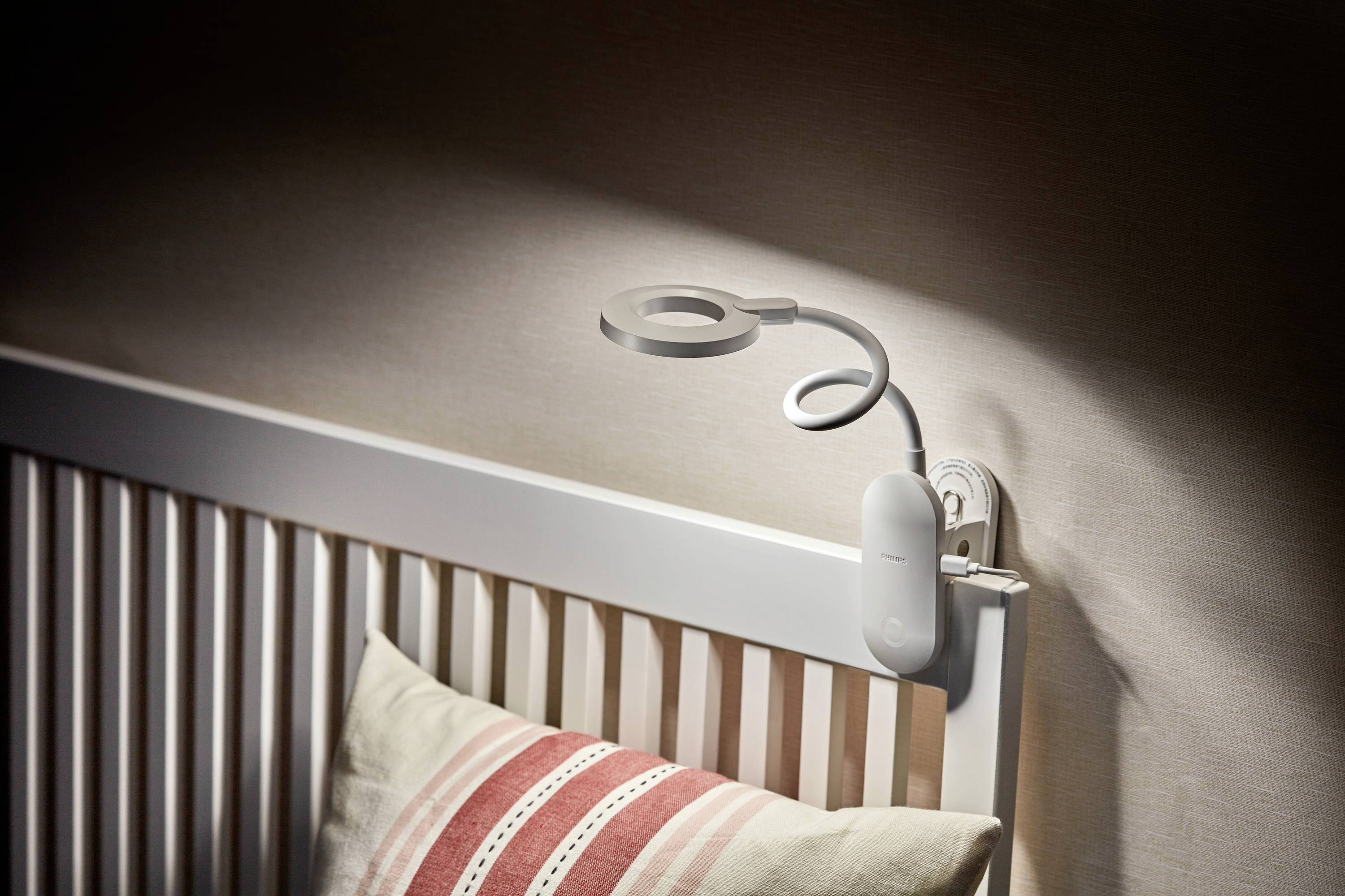 Eine weiße Nachttischlampe mit flexiblem Hals ist an das Kopfende eines weißen Bettes geklemmt. Ein gestreiftes Kissen liegt auf dem Bett.