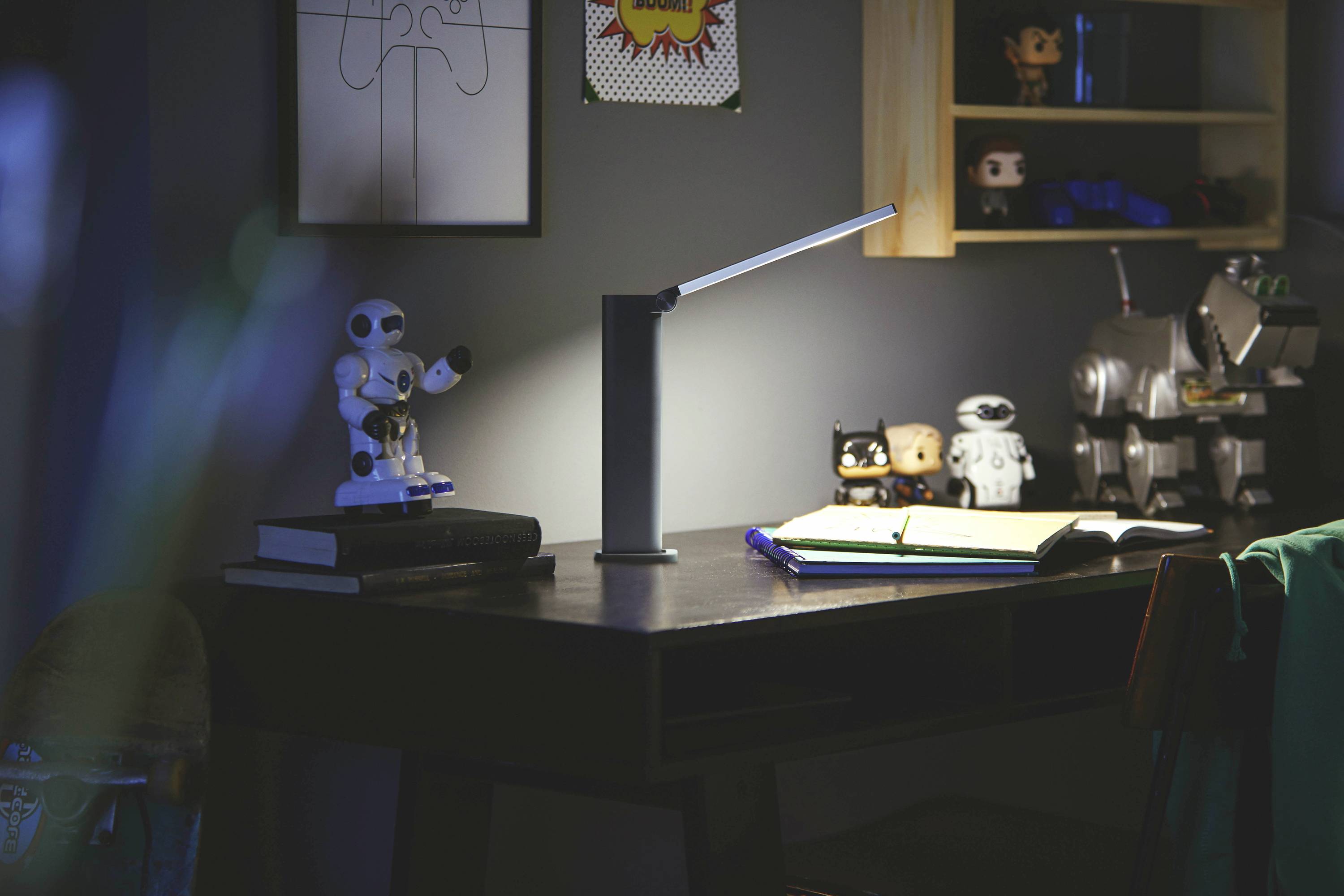 Ein Schreibtisch mit einer eingeschalteten Lampe beleuchtet ein offenes Notizbuch. Spielzeugroboter und Figuren sind im Hintergrund sichtbar.