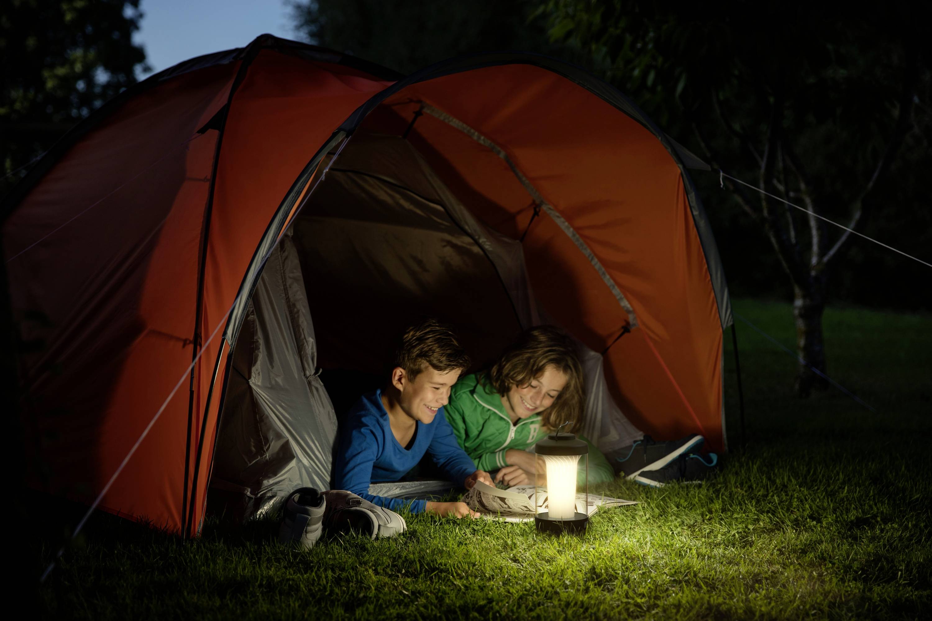 Zwei Kinder liegen abends in einem offenen Zelt auf einer Wiese und lesen bei einer Laterne. Die Szene wirkt friedlich und gemütlich.
