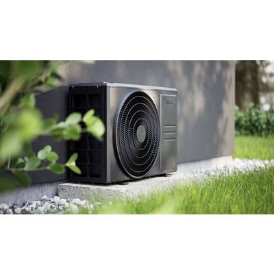 Eine graue Wärmepumpe ist an der Außenwand eines Hauses installiert, umgeben von grünem Gras und Pflanzen im Vordergrund.