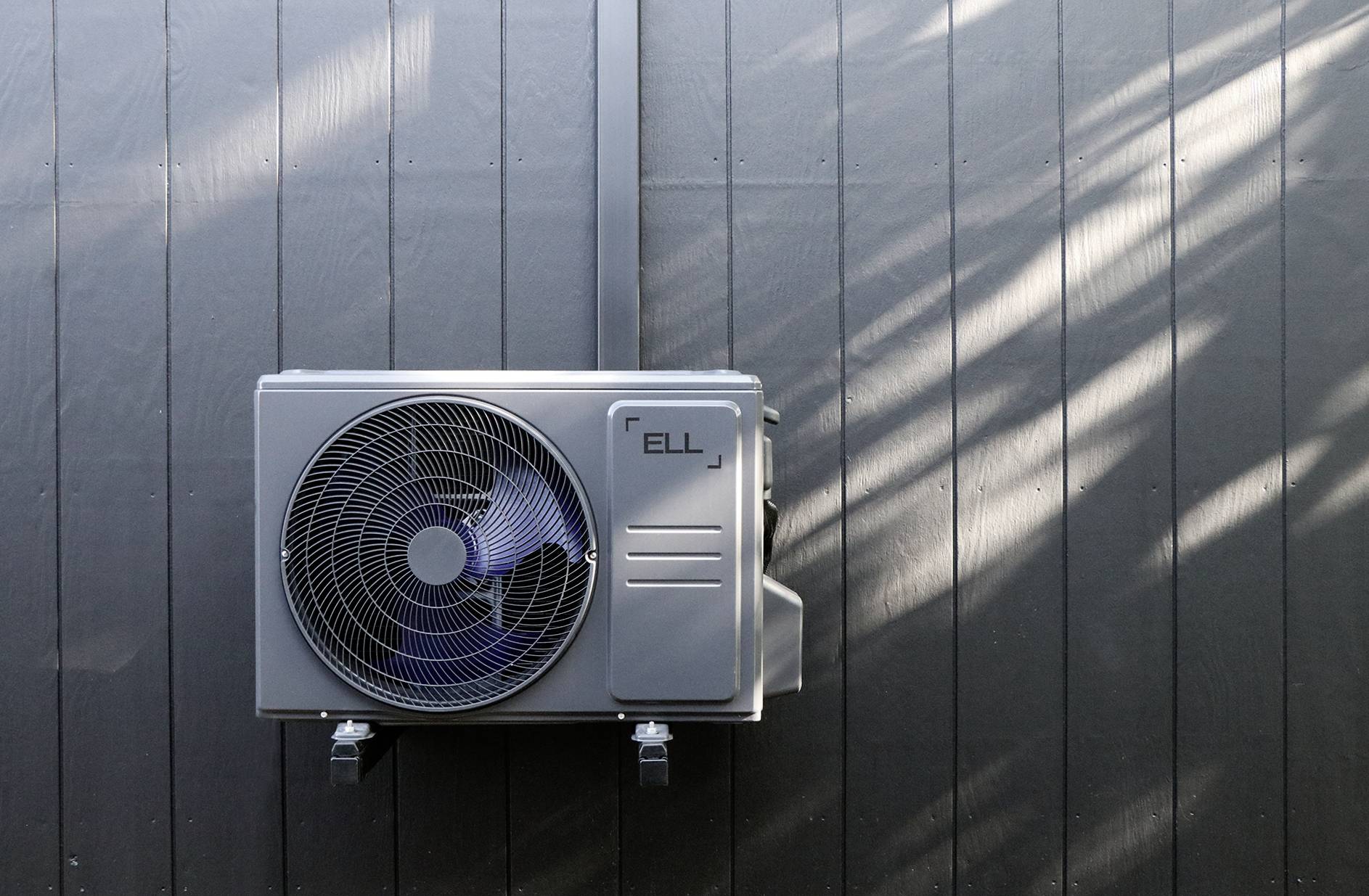 Eine Klimaanlage ist an einer dunklen Wand montiert. Sonnenlicht fällt schräg darauf, und erzeugt leichte Schattenmuster.