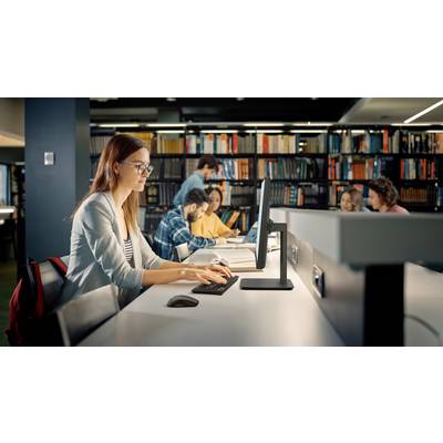 Eine Frau mit Brille arbeitet an einem Computer in einer Bibliothek. Mehrere Personen studieren im Hintergrund, umgeben von Regalen voller Bücher.
