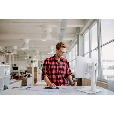 Mann in rotem Karohemd steht an einem Schreibtisch und lächelt auf einen Computerbildschirm in einem hellen, modernen Büro mit großen Fenstern.