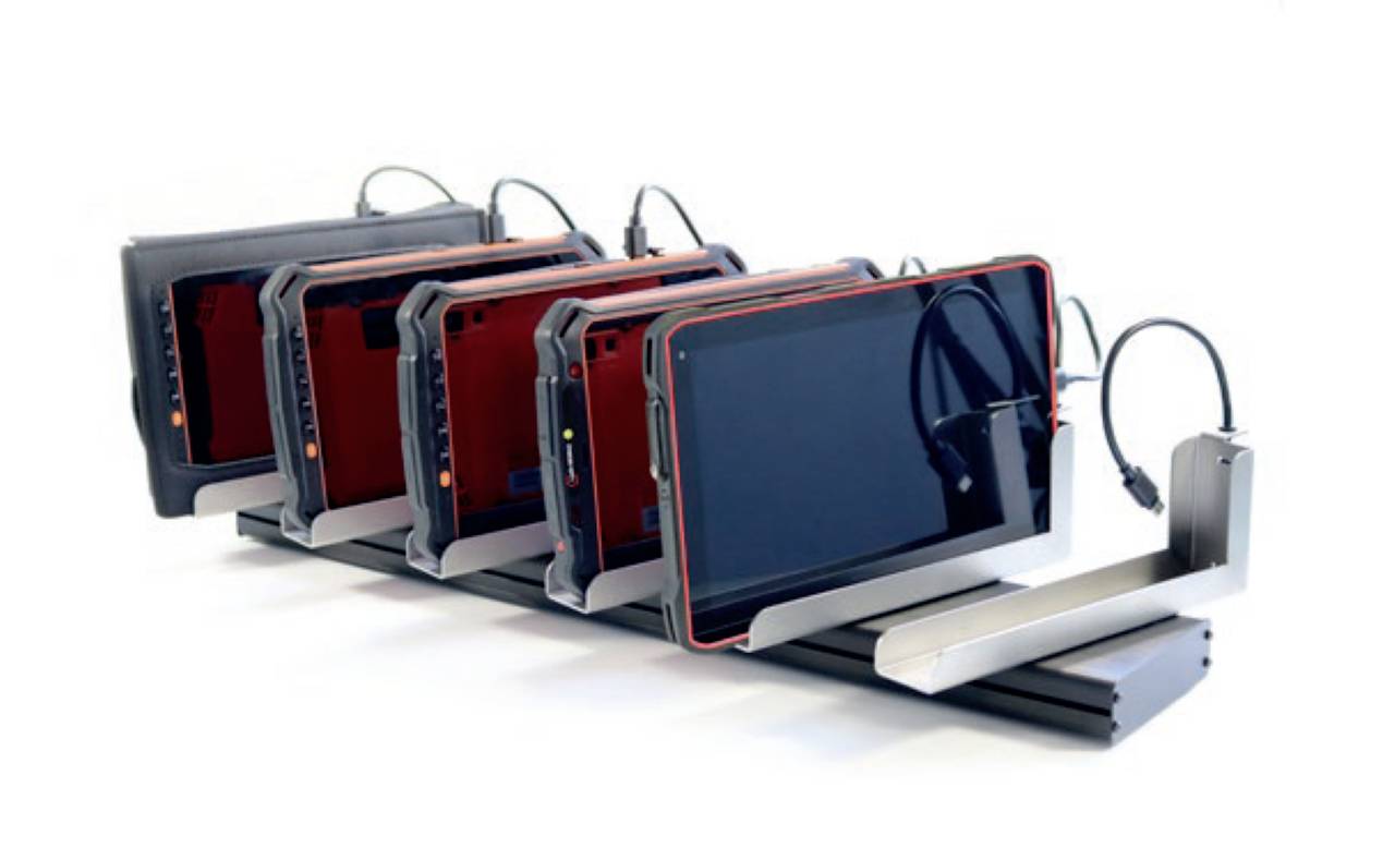 'Vier schwarze Tablets mit rotem Rand stehen nebeneinander in einer Ladestation, verbunden mit Ladekabeln.'