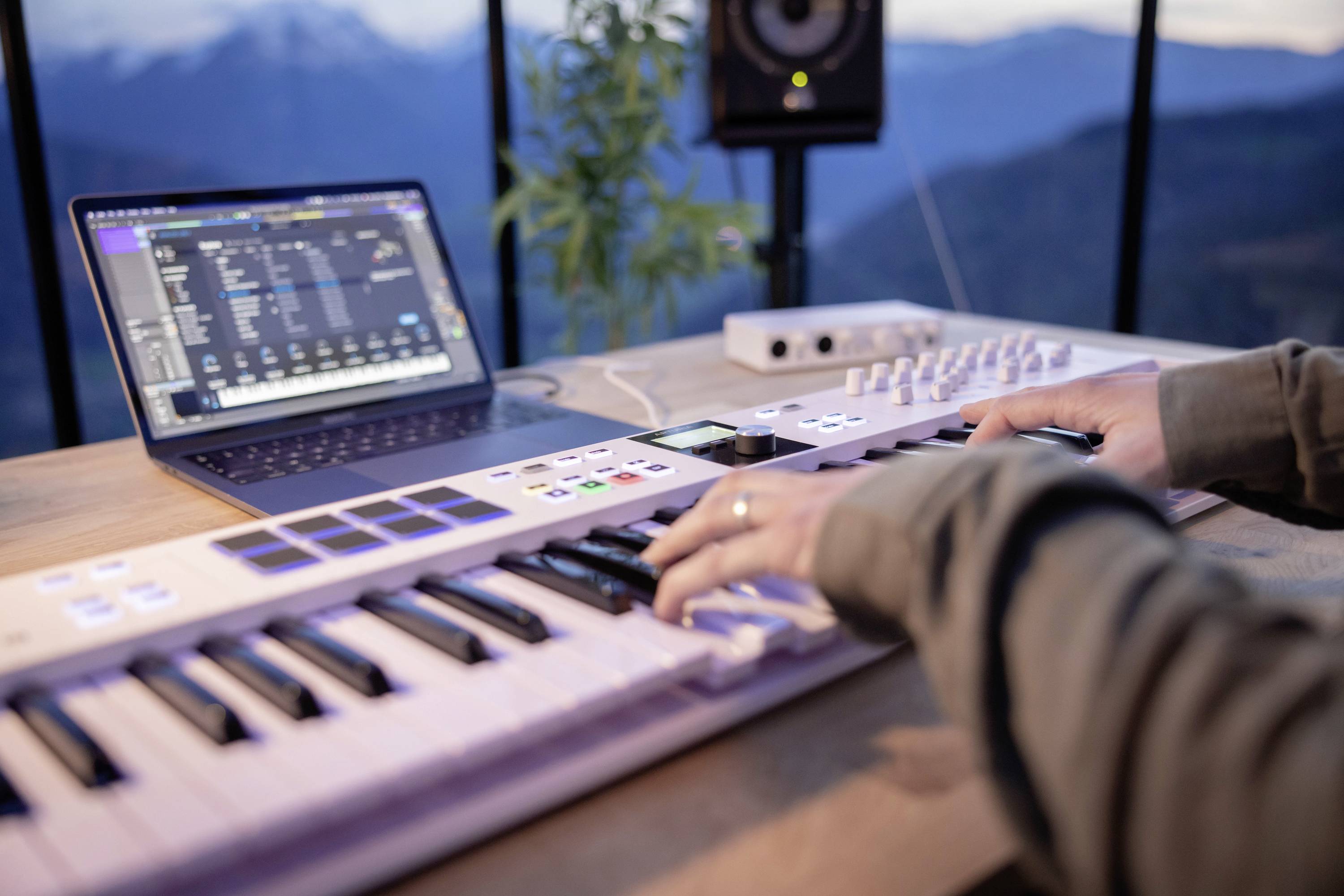 Person spielt auf einem MIDI-Keyboard, verbunden mit einem Laptop, auf dem Musiksoftware läuft. Im Hintergrund eine Berglandschaft.