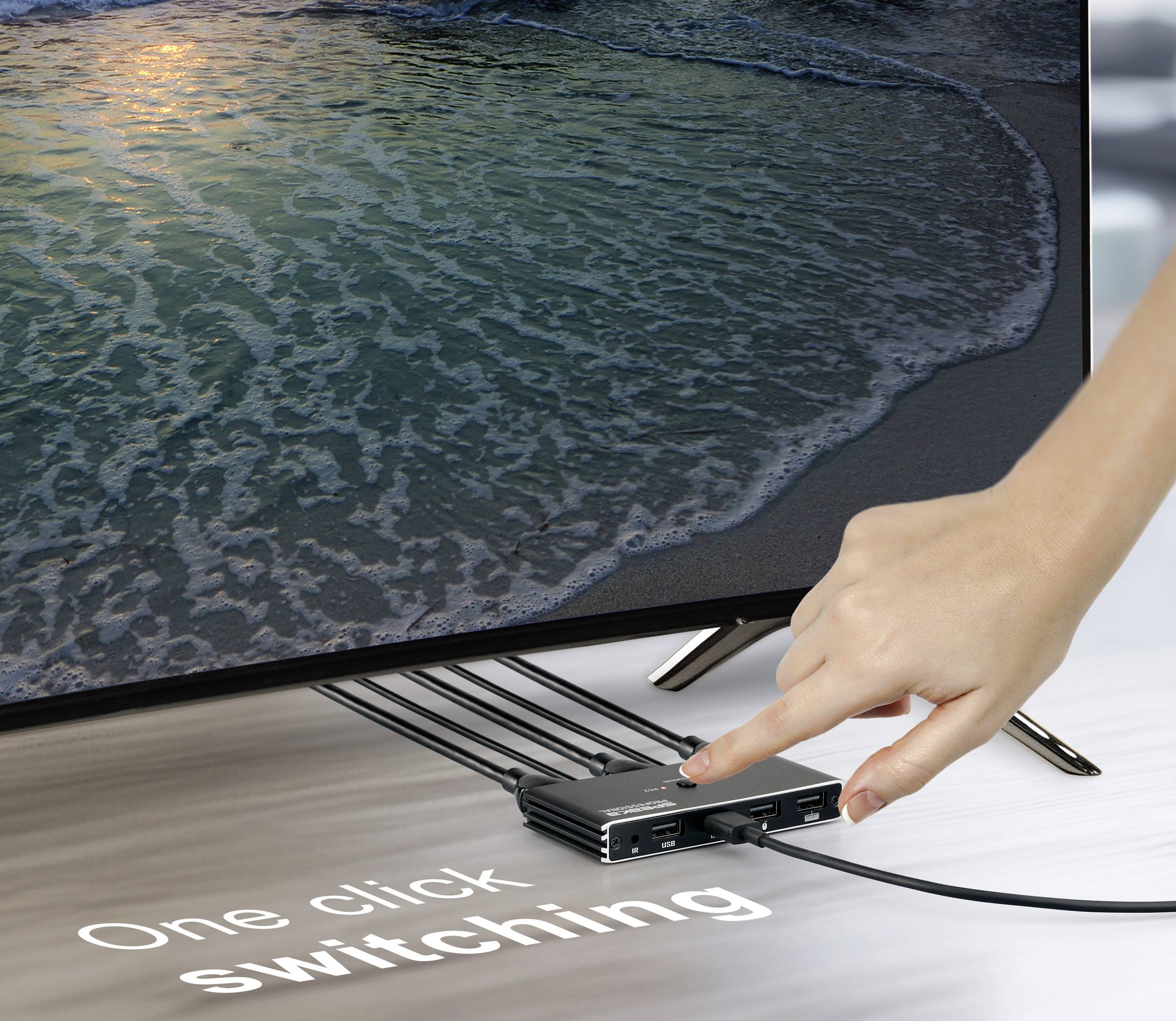 Eine Hand greift nach einem schwarzen Gerät mit Anschlüssen unter einem Fernseher, auf dem eine Strand-Szene zu sehen ist. Der Text lautet „Umschalten mit einem Klick" und deutet auf einfache Bedienbarkeit hin.