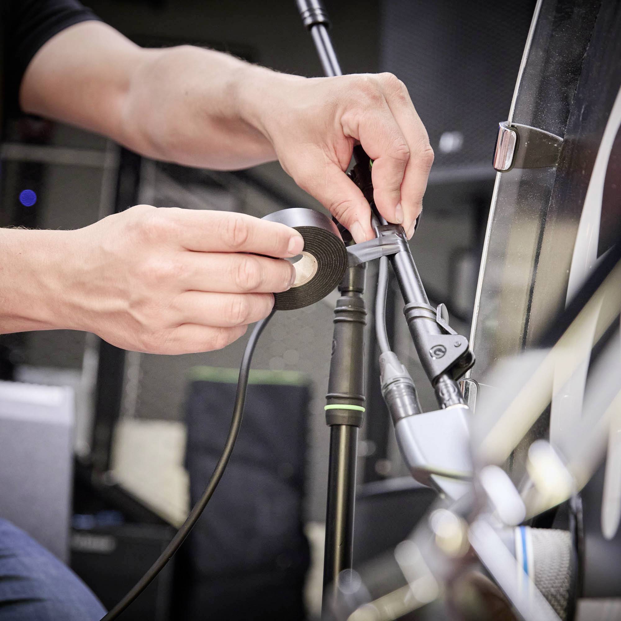 Eine Person befestigt mit Klebeband ein Mikrofonkabel an einem Mikrofonständer. Im Hintergrund sind unscharfe technische Geräte sichtbar.