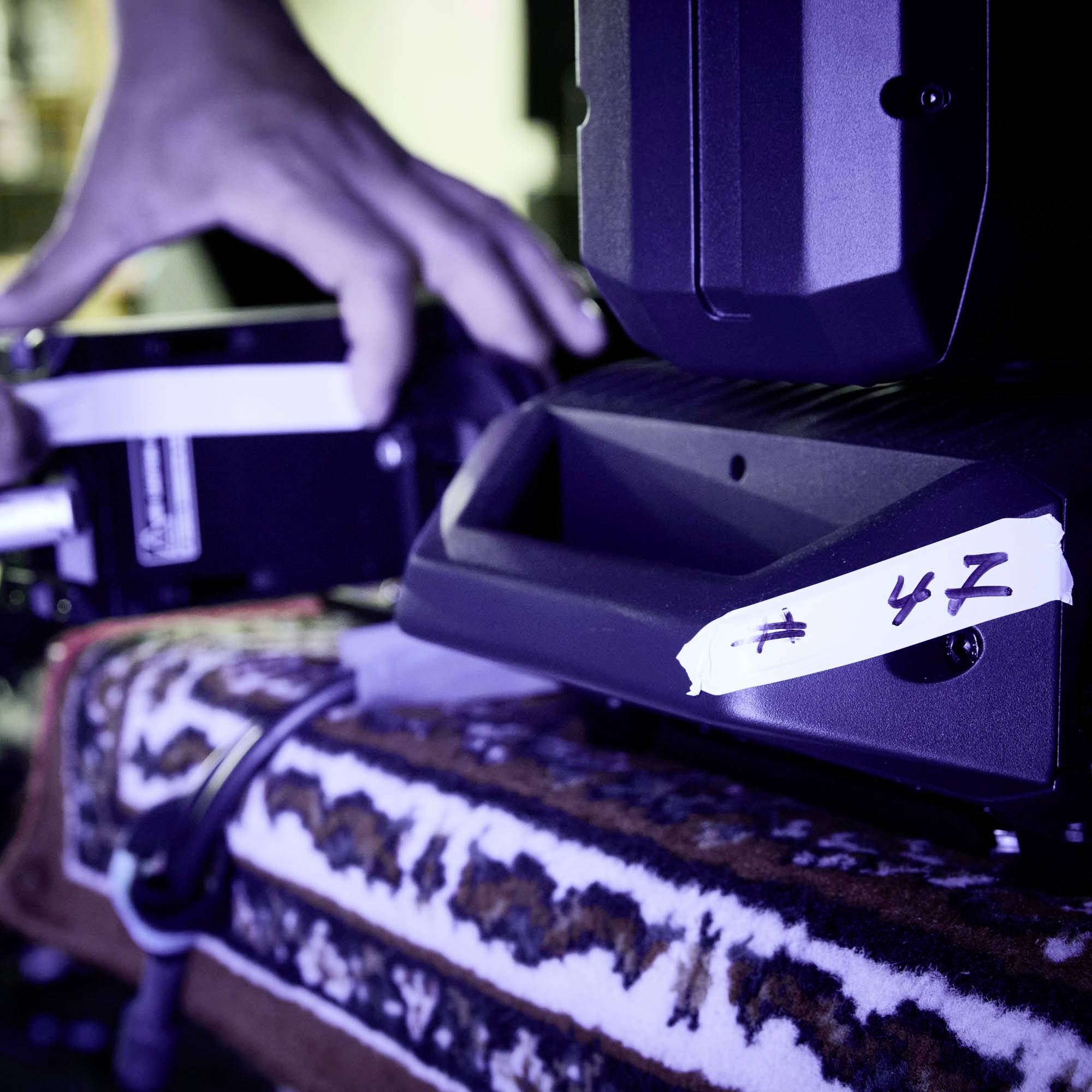 Eine Person justiert ein Kamerastativ in einem abgedunkelten Raum. Auf dem Stativ ist eine beschriftete Nummer 42 sichtbar.