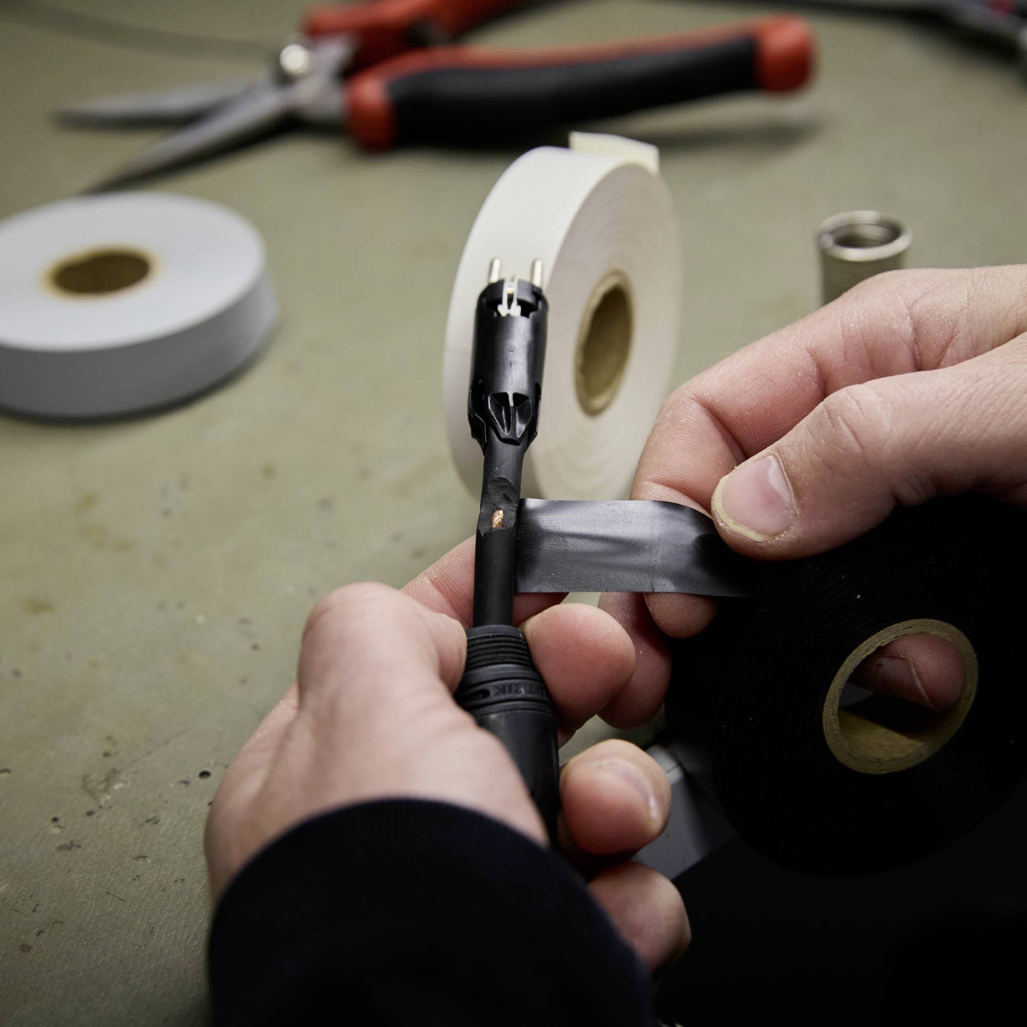 Eine Person isoliert ein Kabel mit schwarzem Klebeband. Im Hintergrund liegen Schere und zwei Klebebandrollen.