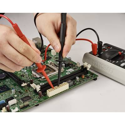 Hände, die Multimeter-Messspitzen verwenden, um eine Leiterplatte zu testen. Die roten und schwarzen Messspitzen sind an verschiedenen Punkten der Platine angeschlossen.