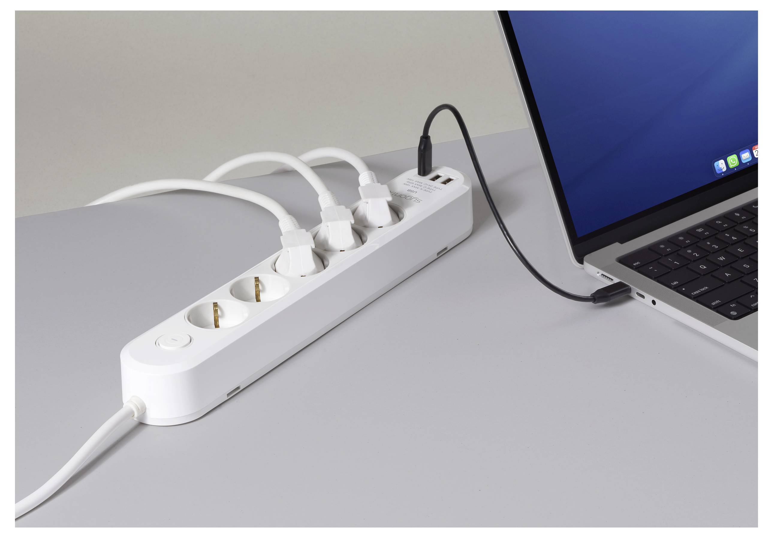 Ein weißer Steckerleiste liegt auf einem Tisch mit drei angeschlossenen Geräten. Ein Laptop, der daneben positioniert ist, ist über ein USB-Kabel verbunden.