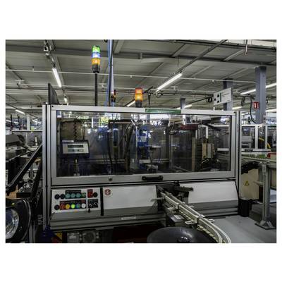 Automatisierte Industriemaschine auf einem Fabrikboden, umschlossen von Sicherheitsglas mit Statusleuchten und Steuertasten, Material auf einem Förderband handhabend.