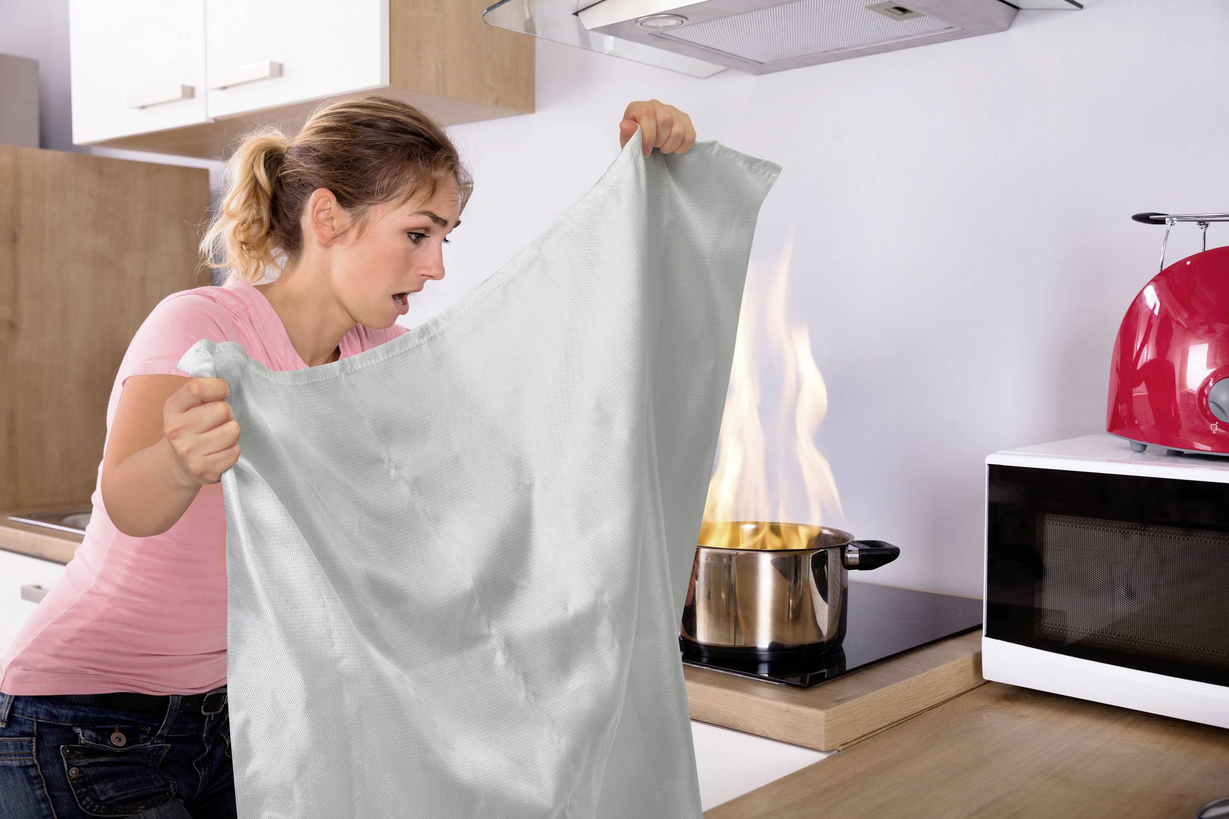 Eine Frau hält eine Decke in der Nähe eines Topfes mit Flammen auf dem Herd und bereitet sich darauf vor, das Feuer zu löschen. Ein Toaster und eine Mikrowelle stehen auf der Theke.