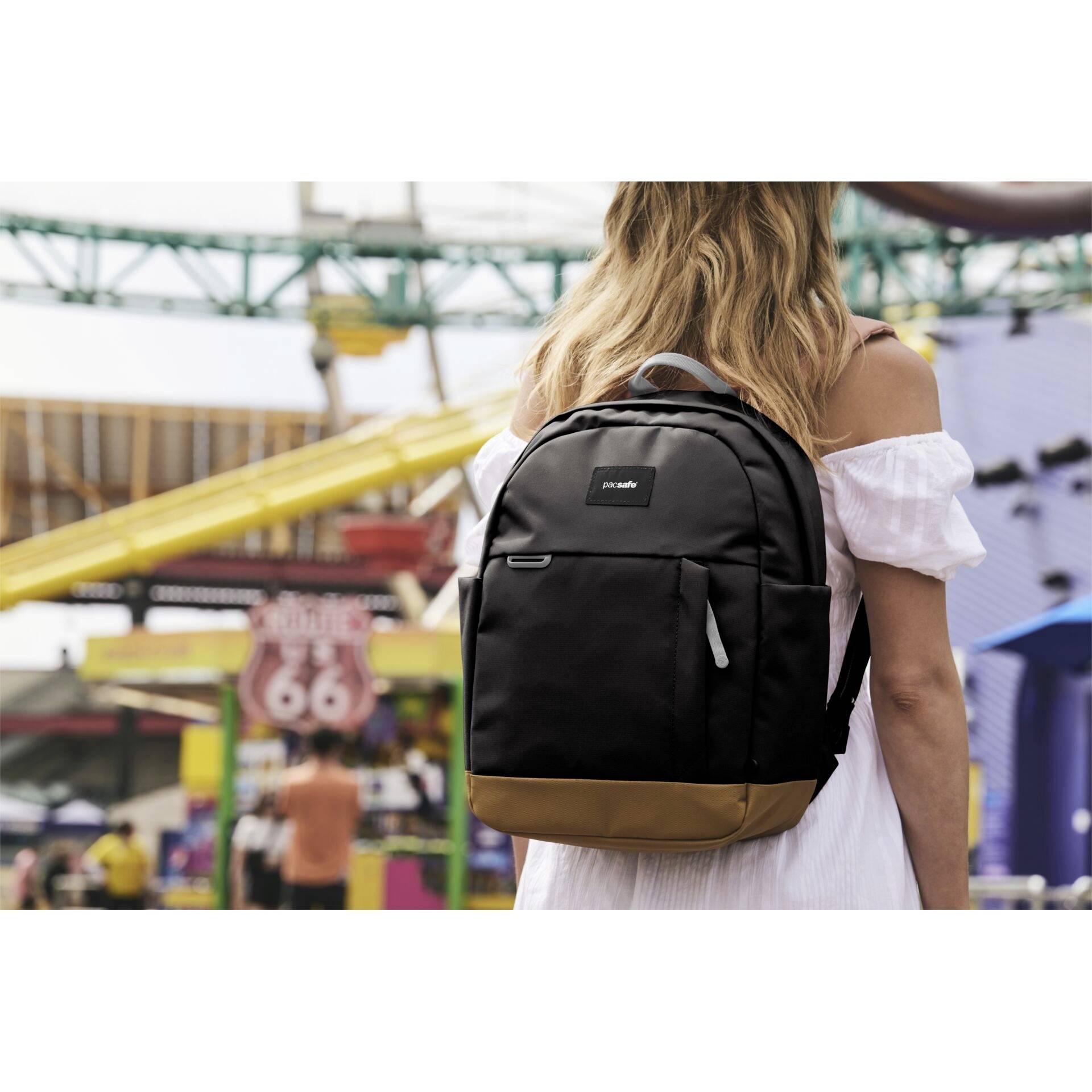 Eine Person mit langen Haaren steht im Freien und trägt einen schwarz-braunen Rucksack, während im Hintergrund bunte Fahrgeschäfte eines Vergnügungsparks zu sehen sind.