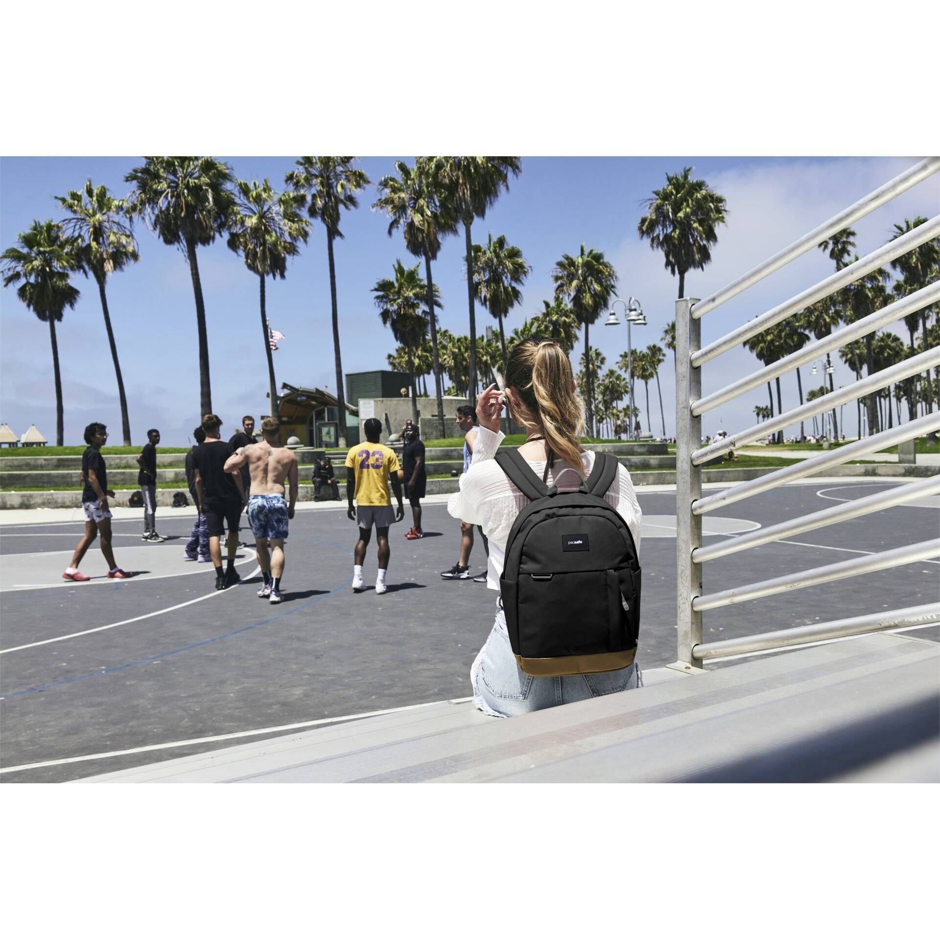 Eine Person mit einem Rucksack sitzt auf den Zuschauerrängen und beobachtet eine Gruppe, die Basketball auf einem Freiplatz spielt, während im Hintergrund Palmen stehen.