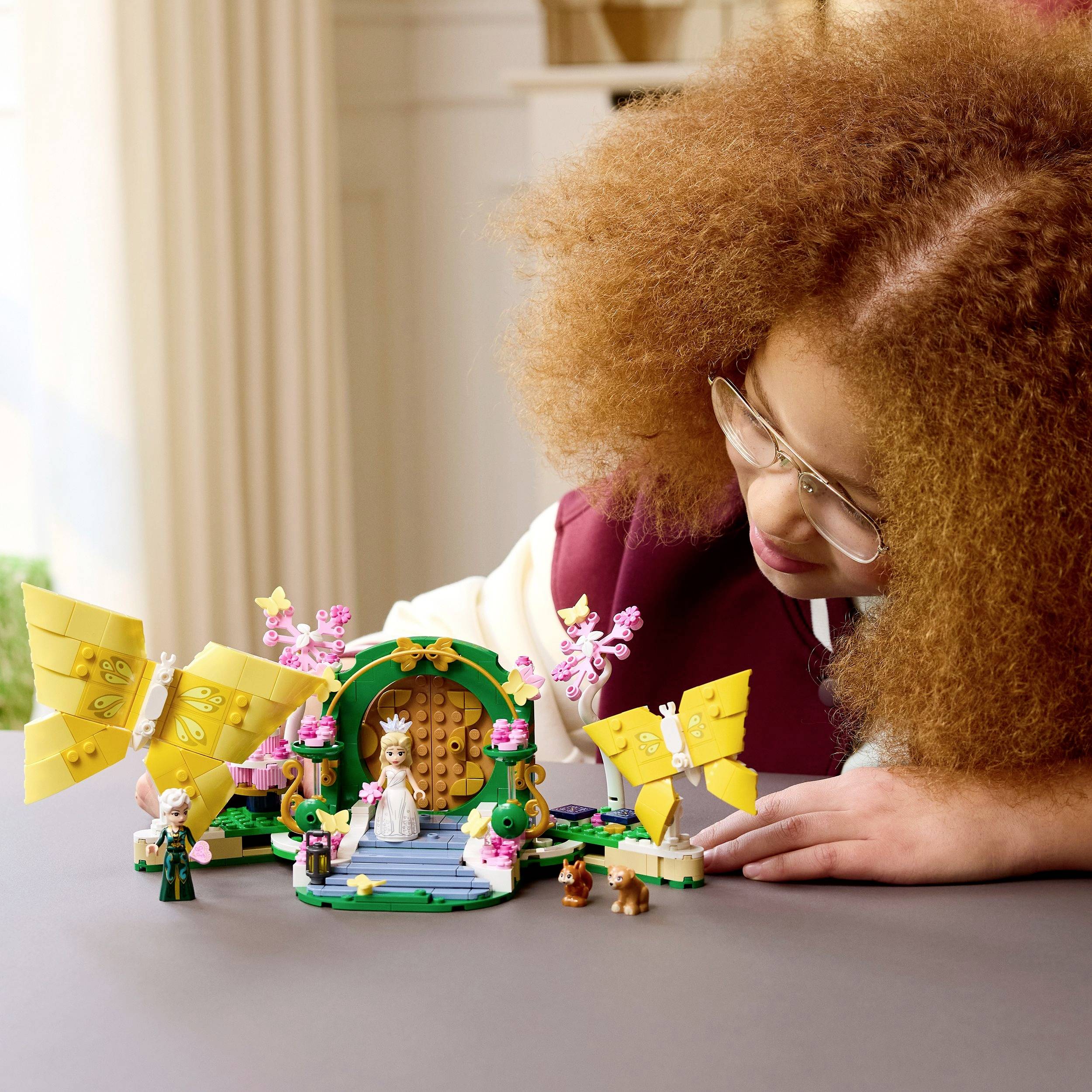 Ein Kind mit lockigem Haar und Brille spielt mit einem Miniatur-Spielzeugset, das eine Burg, Figuren und Bäume auf einem Tisch im Innenraum umfasst.