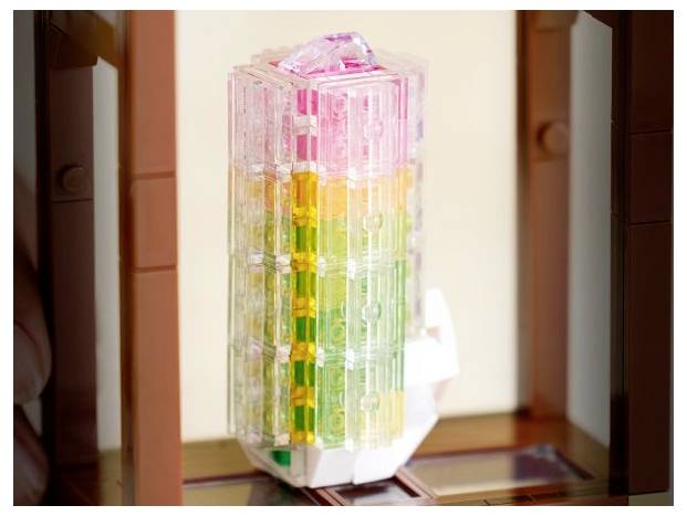 Bunter durchscheinender Turm aus Klötzen in einem braunen Rahmen, der rosa, gelbe, grüne und weiße Abschnitte aufweist. Scheint aus Plastik gefertigt zu sein.