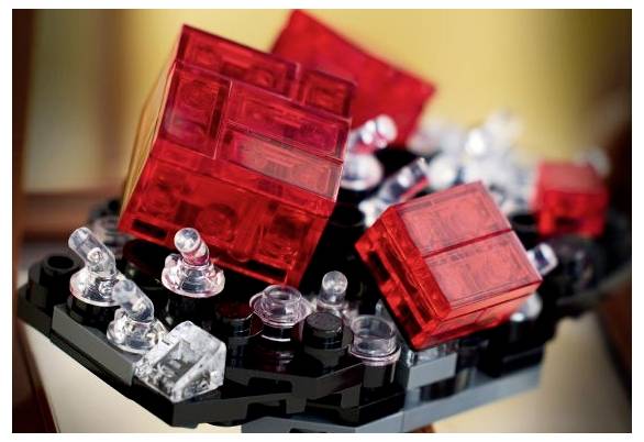 Rote und durchsichtige Lego-Steine auf einer schwarzen Grundfläche angeordnet, die eine abstrakte, geometrische Form mit schimmernden Lichtreflexen bilden.