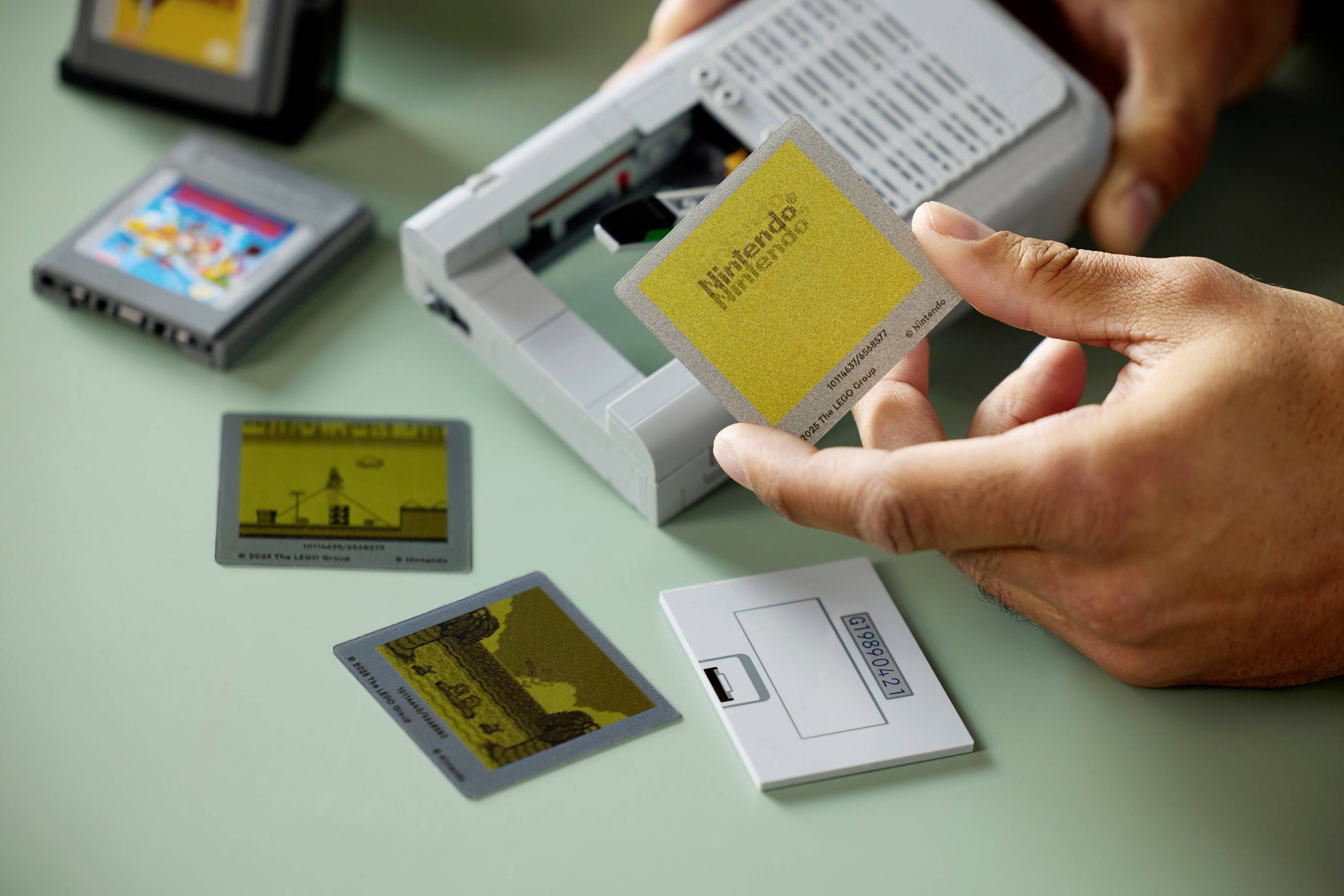 Eine Person steckt eine Spielekarte in eine Retro-Handheld-Spielekonsole, umgeben von mehreren anderen Karten mit Spieletiteln, auf einem Tisch.