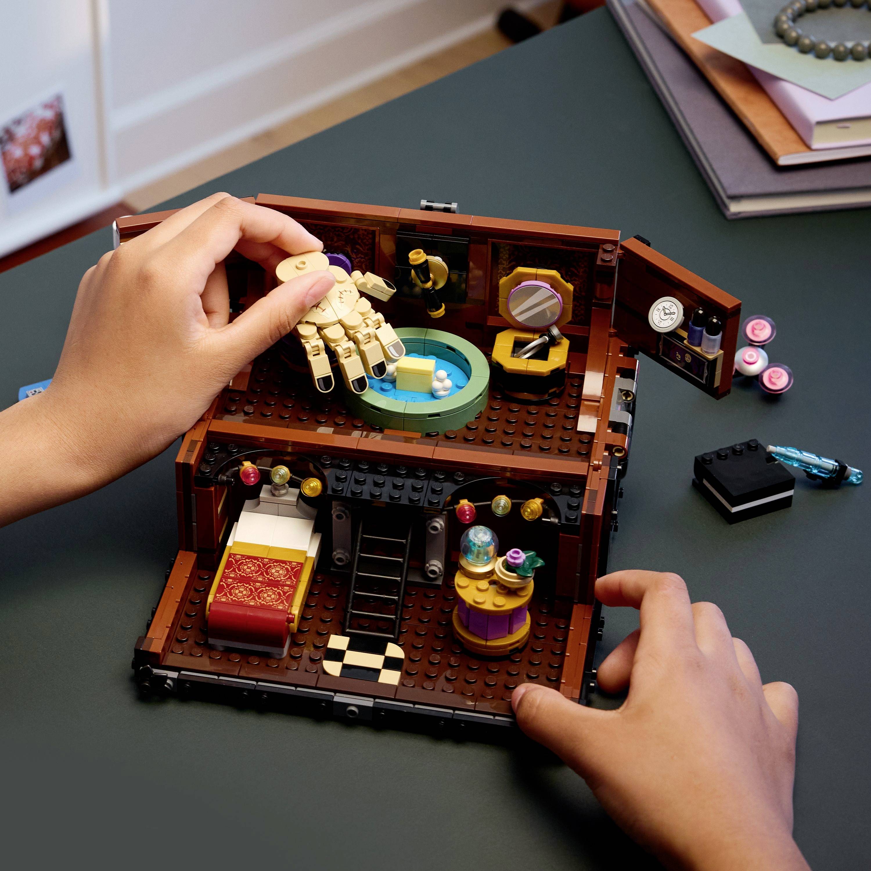 Zwei Hände interagieren mit einem miniaturhaften, geöffneten LEGO-Puppenhaus. Eine Hand positioniert eine LEGO-Figur in einer Badewanne; die andere hält das Haus stabil.