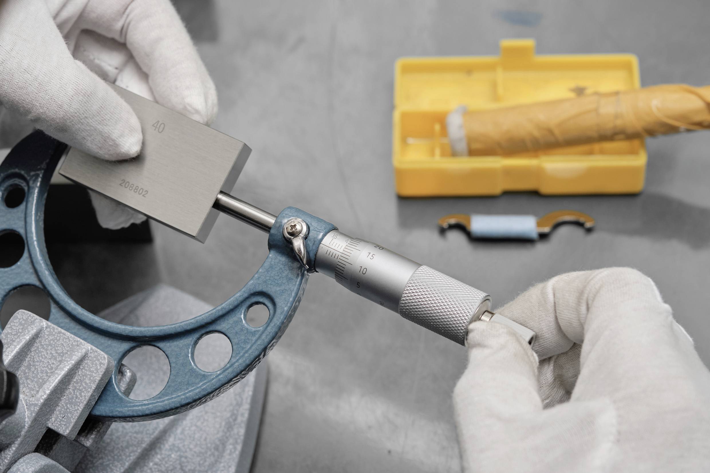 Eine Person mit Handschuhen verwendet ein Messgerät (Mikrometer), um einen Metallblock auf einer Werkbank zu vermessen, während im Hintergrund ein gelber Werkzeugkoffer zu sehen ist.