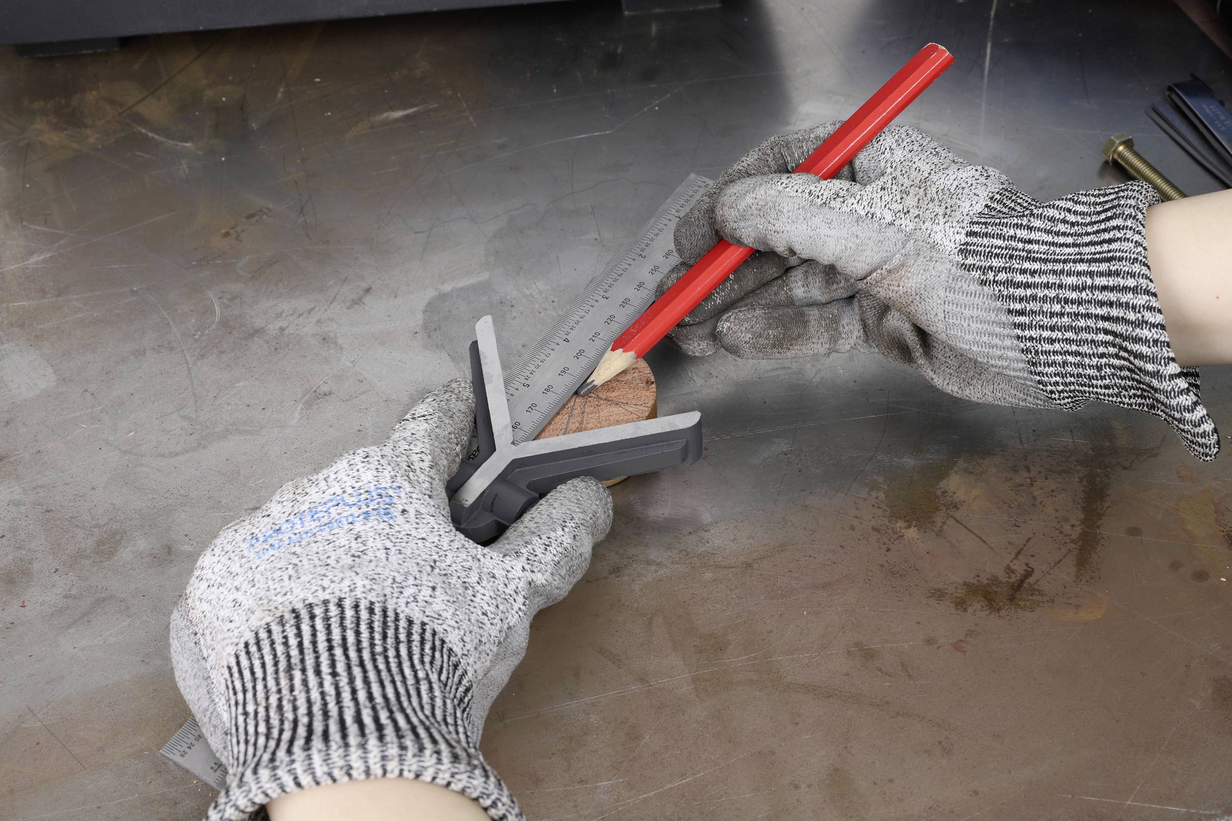 Eine Person mit Sicherheitshandschuhen verwendet ein Winkelmaß und einen roten Bleistift, um ein Holzstück auf einer Metalloberfläche zu markieren.