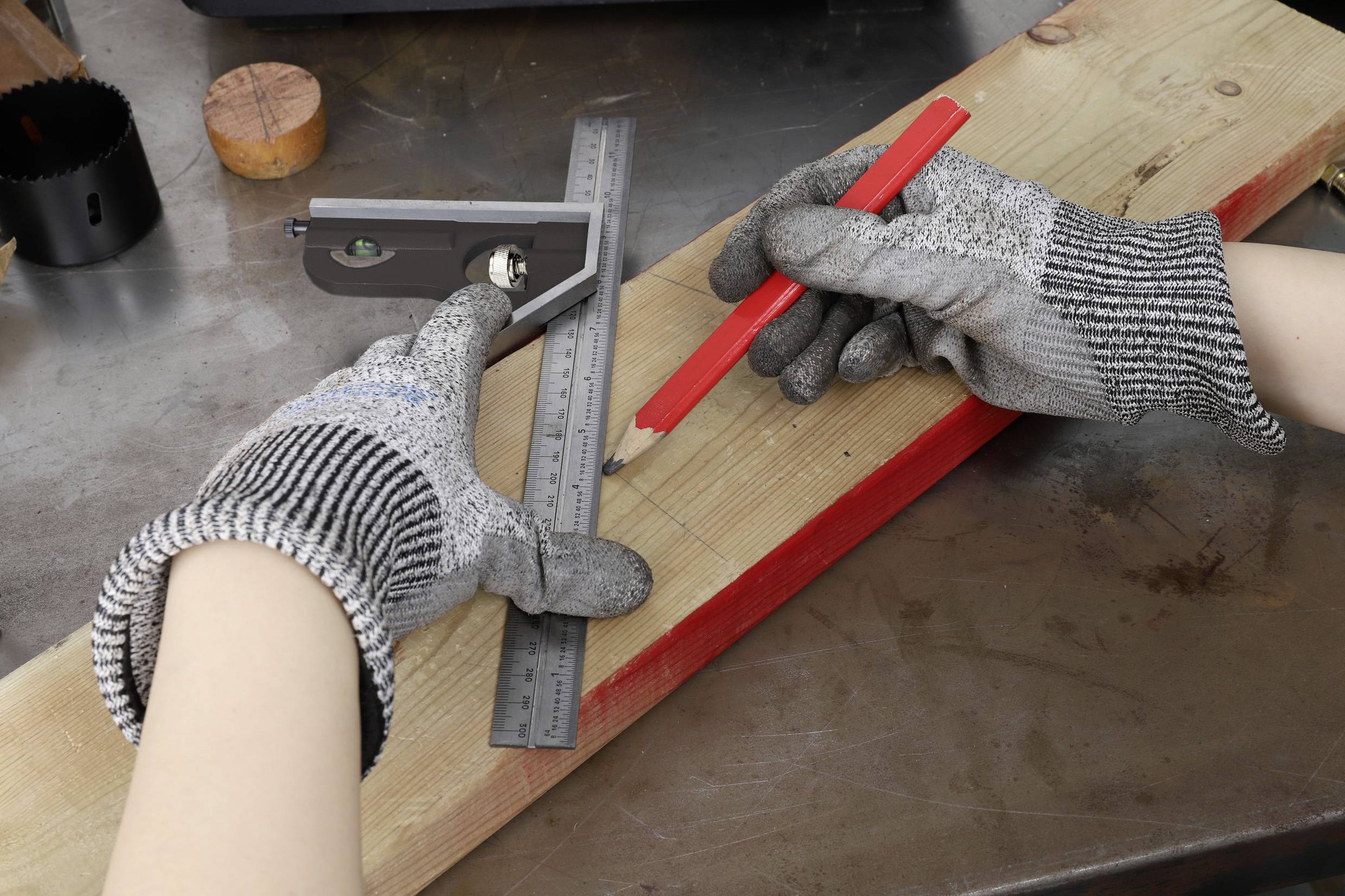 Eine Person mit Handschuhen misst und markiert ein Holzbrett mit einem Lineal und einem roten Bleistift auf einer Metalloberfläche.