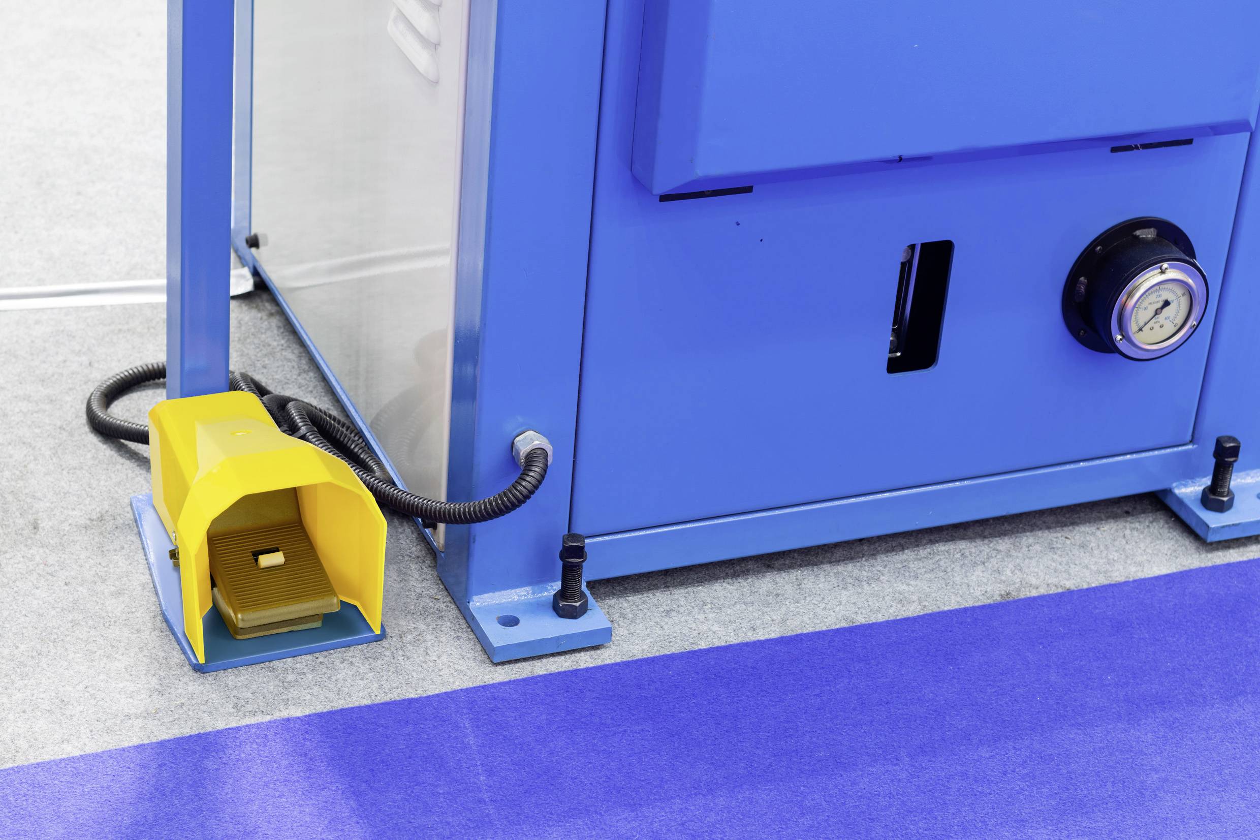 Eine blaue Industriemaschine auf einem grauen Boden mit einer lila Matte in der Nähe. Ein gelbes Fußpedal ist seitlich positioniert.