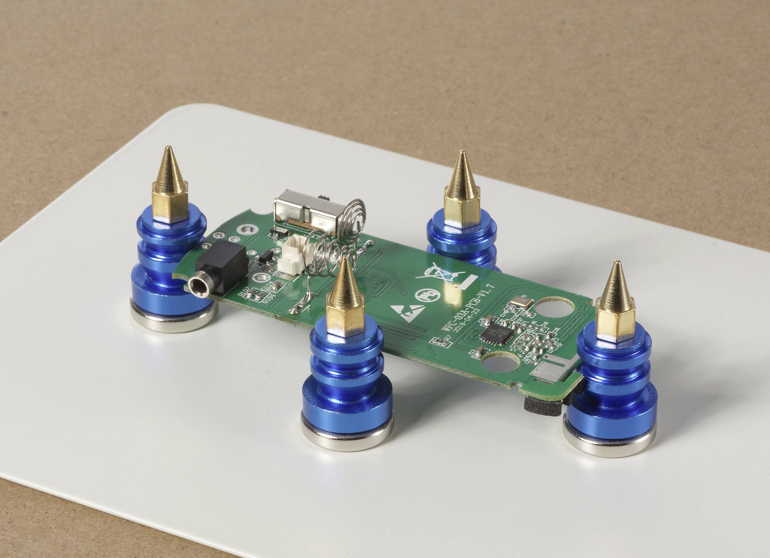 Eine grüne Leiterplatte mit blauen und goldenen Abstandshaltern liegt auf einer weißen Oberfläche und zeigt Komponenten und Anschlüsse für die elektronische Montage.