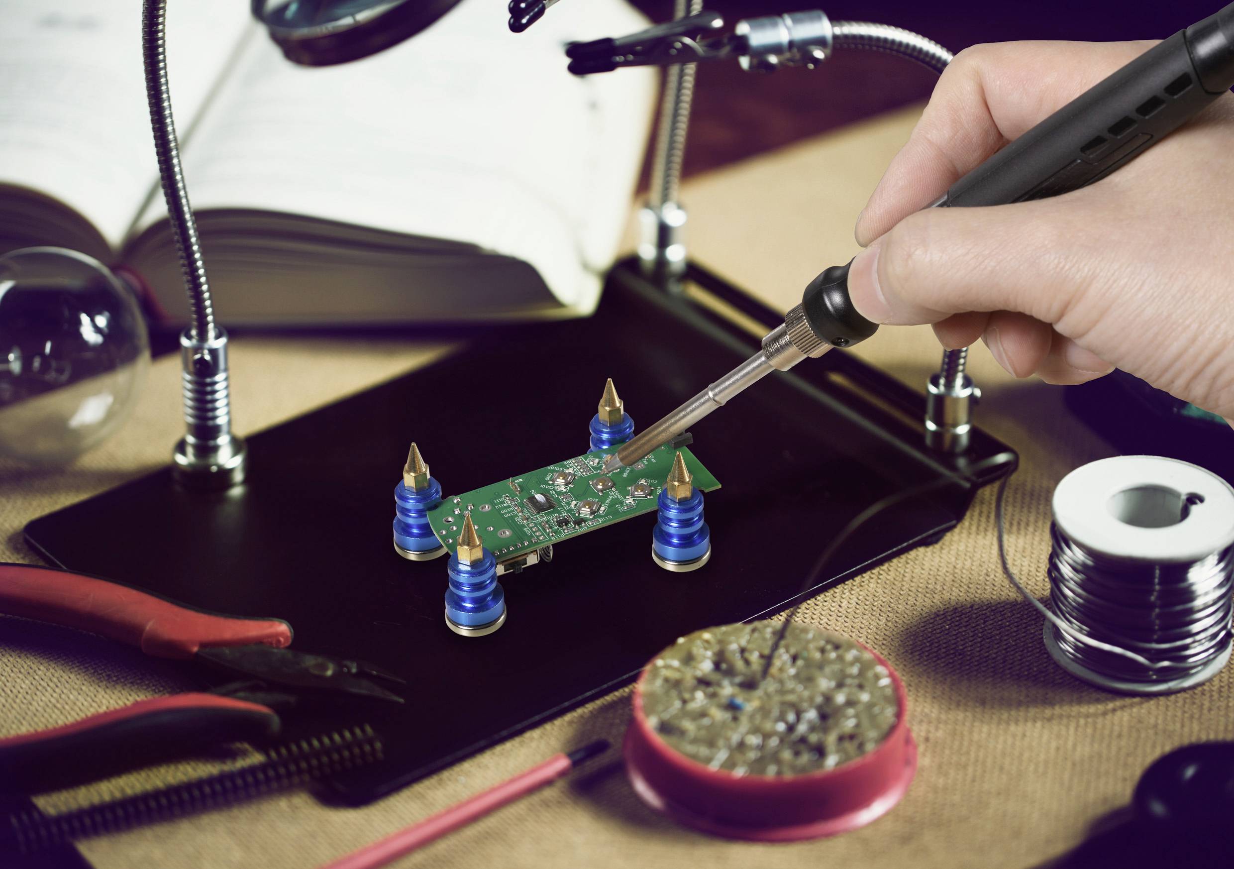 Eine Person, die einen Lötkolben verwendet, um an einer kleinen grünen Leiterplatte zu arbeiten, die auf einem Ständer befestigt ist, umgeben von Werkzeugen und Kabeln.