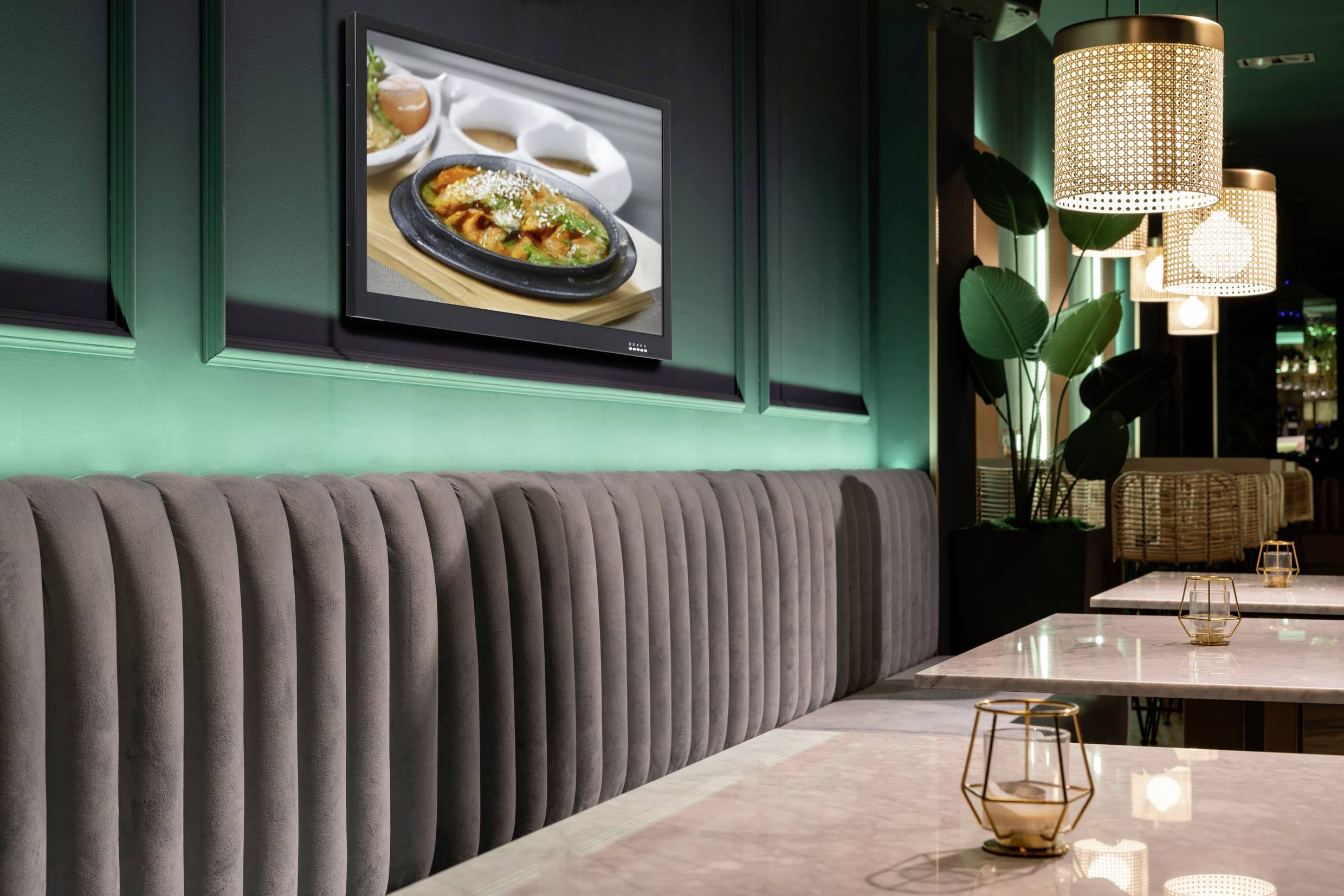 Modernes Restaurantinterieur mit gemütlichen grauen Sitzgelegenheiten, einem an der Wand montierten Fernseher, der ein Gericht zeigt, warmem Licht und eleganten Tischen.