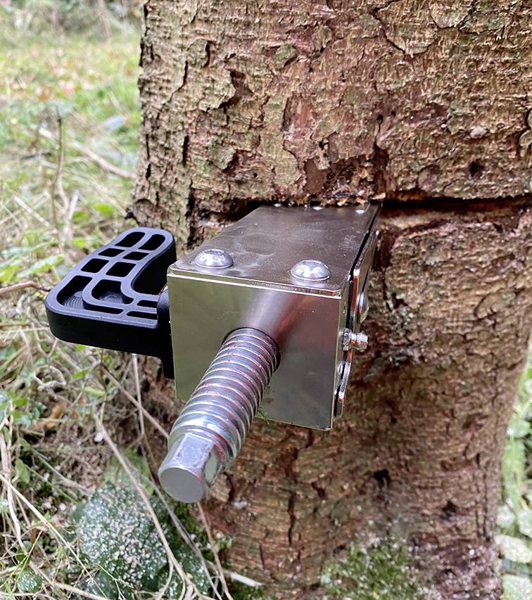 Ein Metallwerkzeug wird in einen Baumstamm eingeschlagen. Es scheint ein Spaltkeilen mit einem schwarzen Griff zu sein, der in einem bewaldeten Gebiet platziert ist.