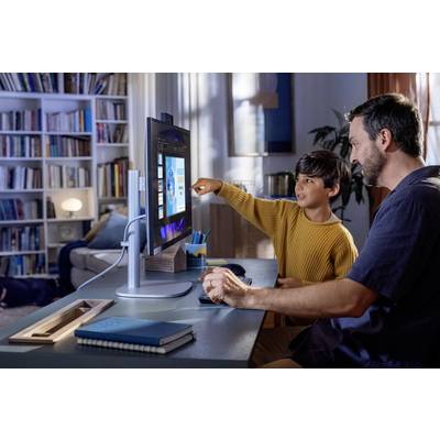 Ein Vater und Sohn sitzen an einem Schreibtisch und konzentrieren sich auf einen Computerbildschirm. Der Sohn zeigt auf den Bildschirm, während der Vater die Maus bedient. Bücherregale im Hintergrund deuten auf ein Home Office hin.