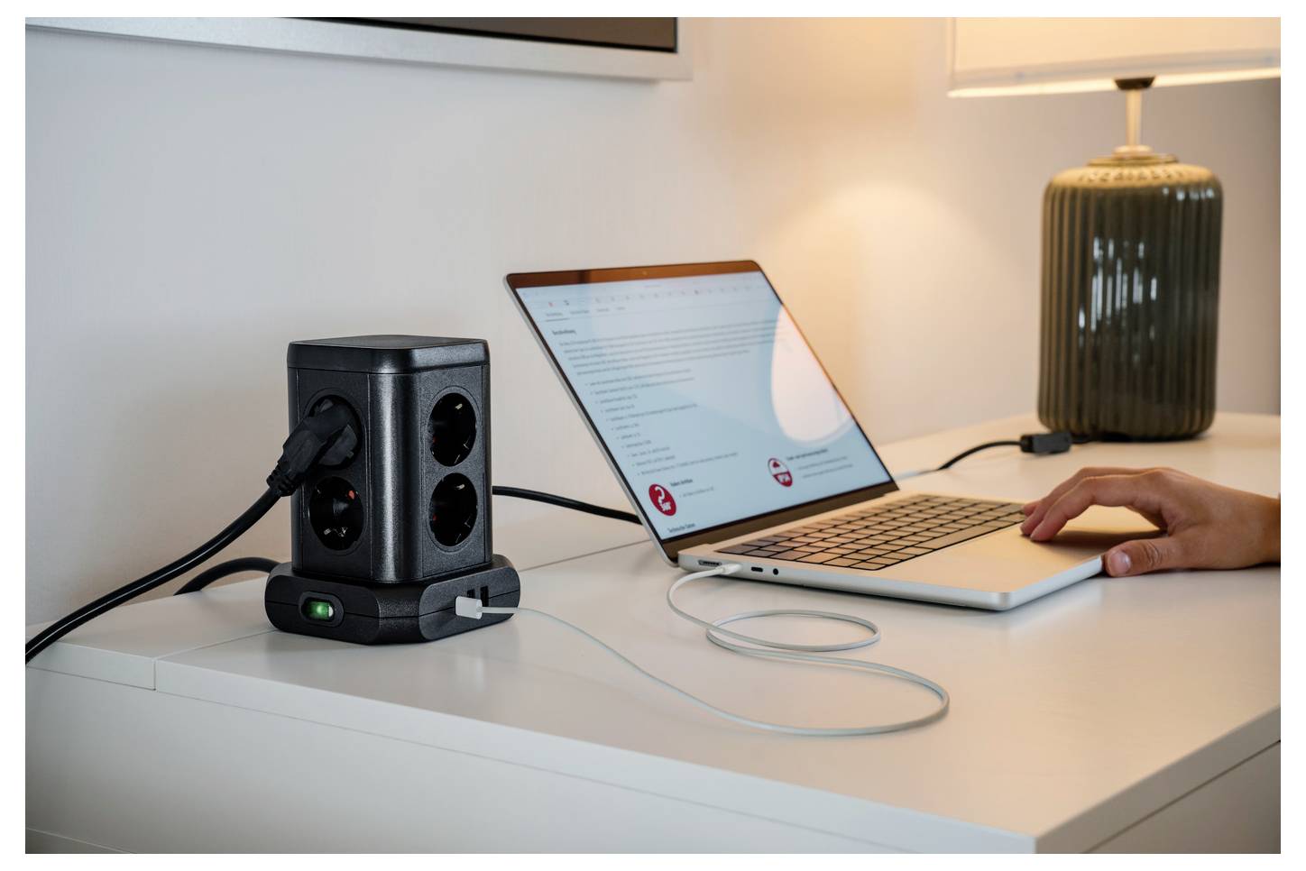 Ein Laptop lädt auf einem Schreibtisch von einer schwarzen Stromstation auf, mit einer Hand auf der Tastatur und einer leuchtenden Tischlampe im Hintergrund.