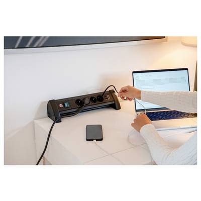 Eine Person steckt ein Kabel in eine Steckdosenleiste auf einem Schreibtisch neben einem Laptop und einer externen Festplatte auf einem weißen Tisch.