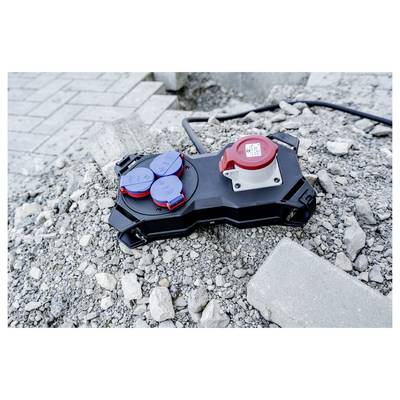 Tragbare Stromverteilungsbox mit roten und blauen Steckdosen liegt auf Kies und Bauschutt neben einer gepflasterten Oberfläche.