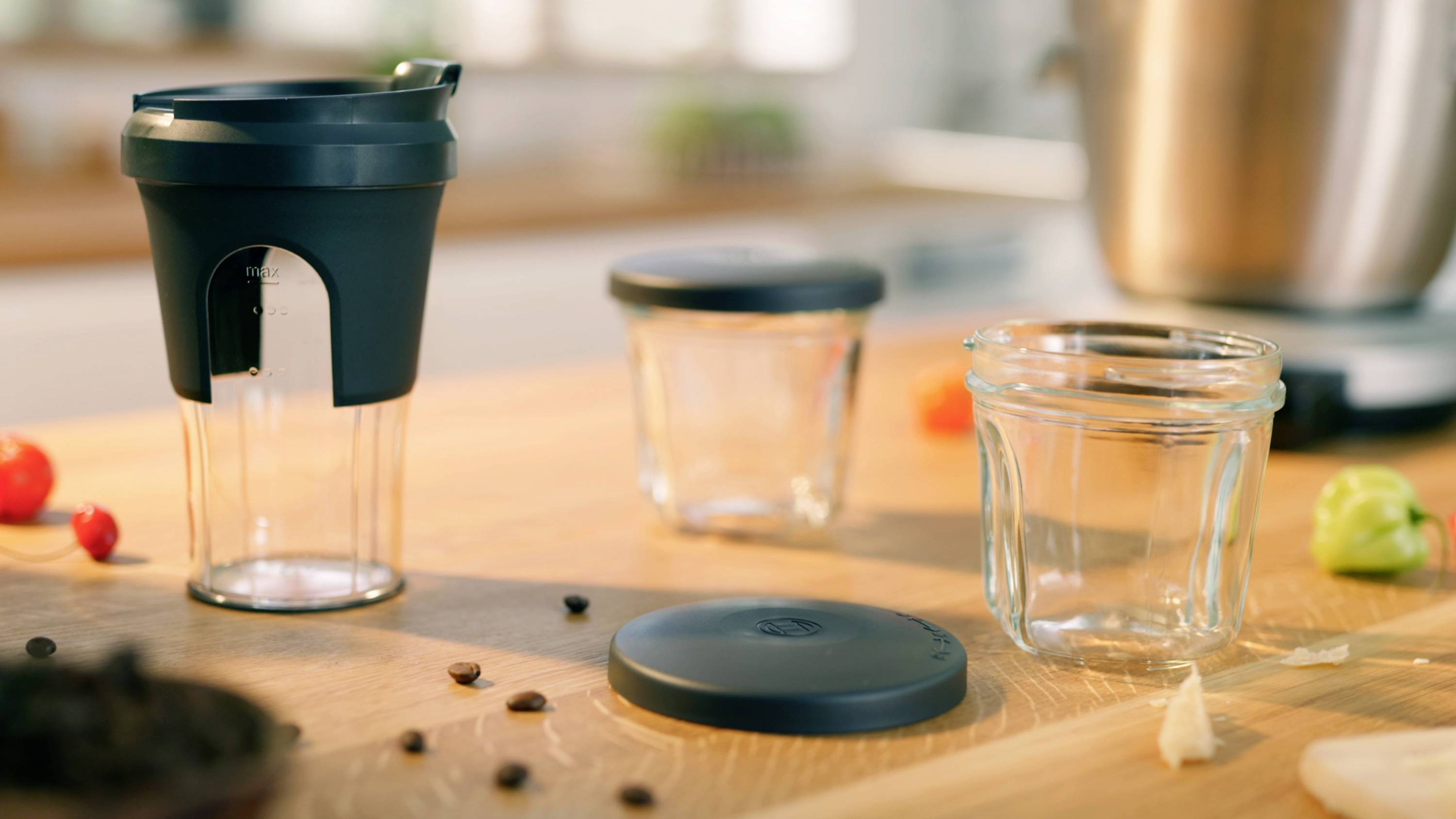 Eine Küchenarbeitsplatte mit Glasgefäßen, Deckeln und einem schwarzen Kaffeemacher. Verstreute Kaffeebohnen, Kirschtomaten und Knoblauch sind zu sehen.