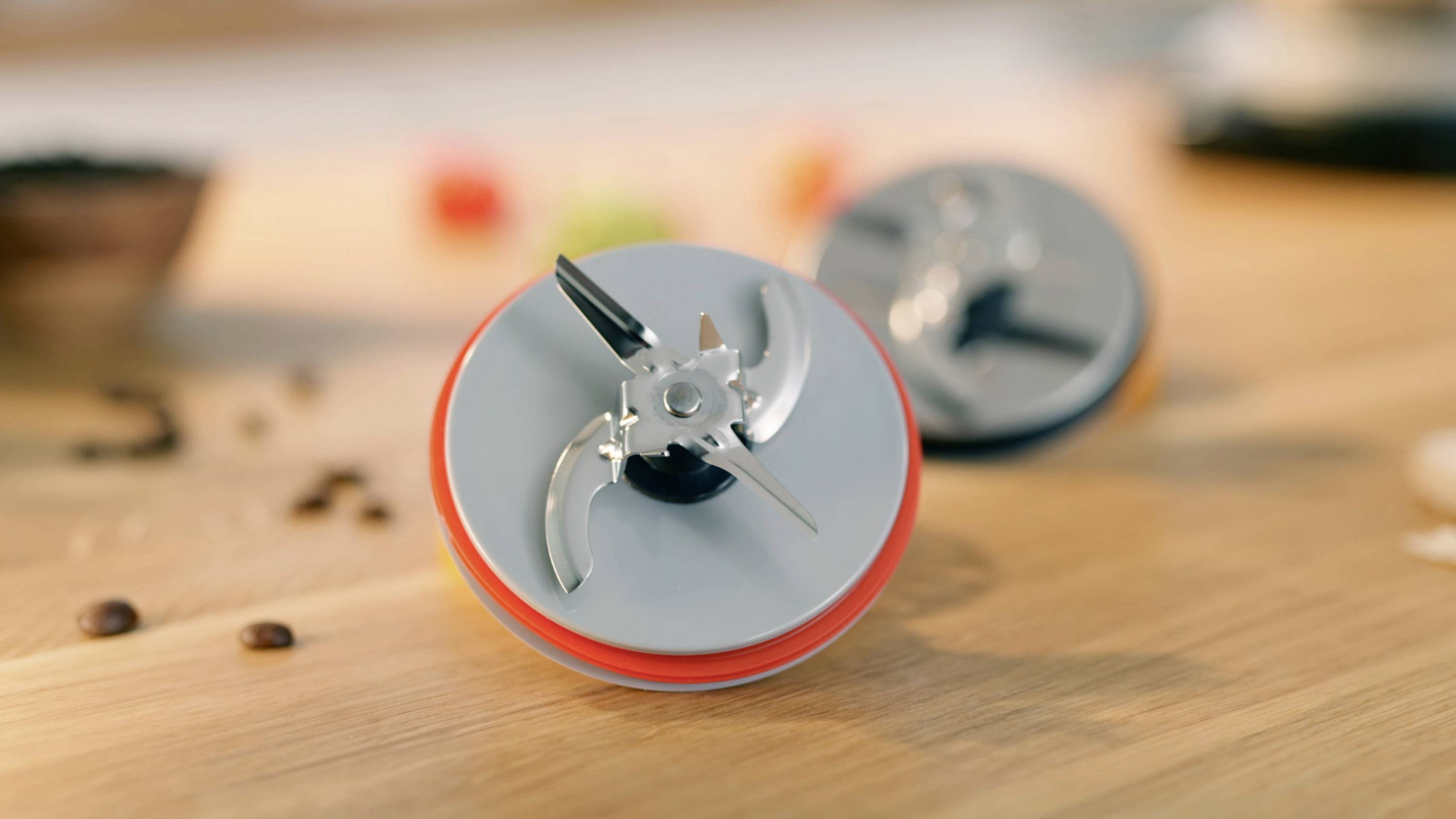Zwei Mixer-Klingen mit scharfen, silbernen Kanten liegen auf einer Holzoberfläche, umgeben von einigen verstreuten Kaffeebohnen.