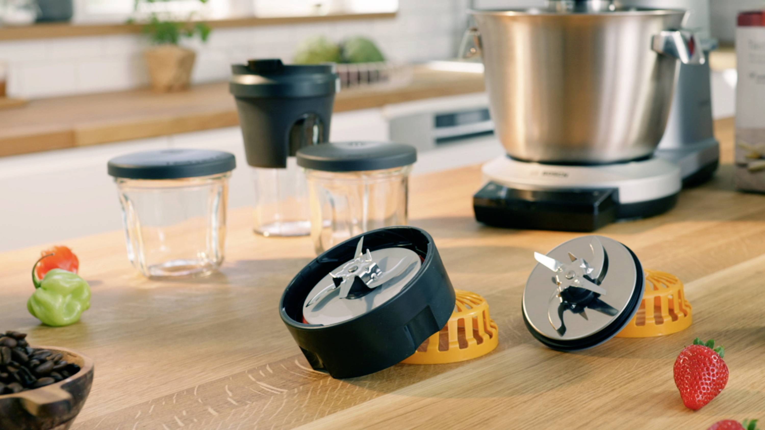 Mixer-Zubehör auf einer Küchenarbeitsplatte mit einer Rührschüssel im Hintergrund, die Klingen, Behälter und frische Zutaten wie Paprika und eine Erdbeere präsentiert.