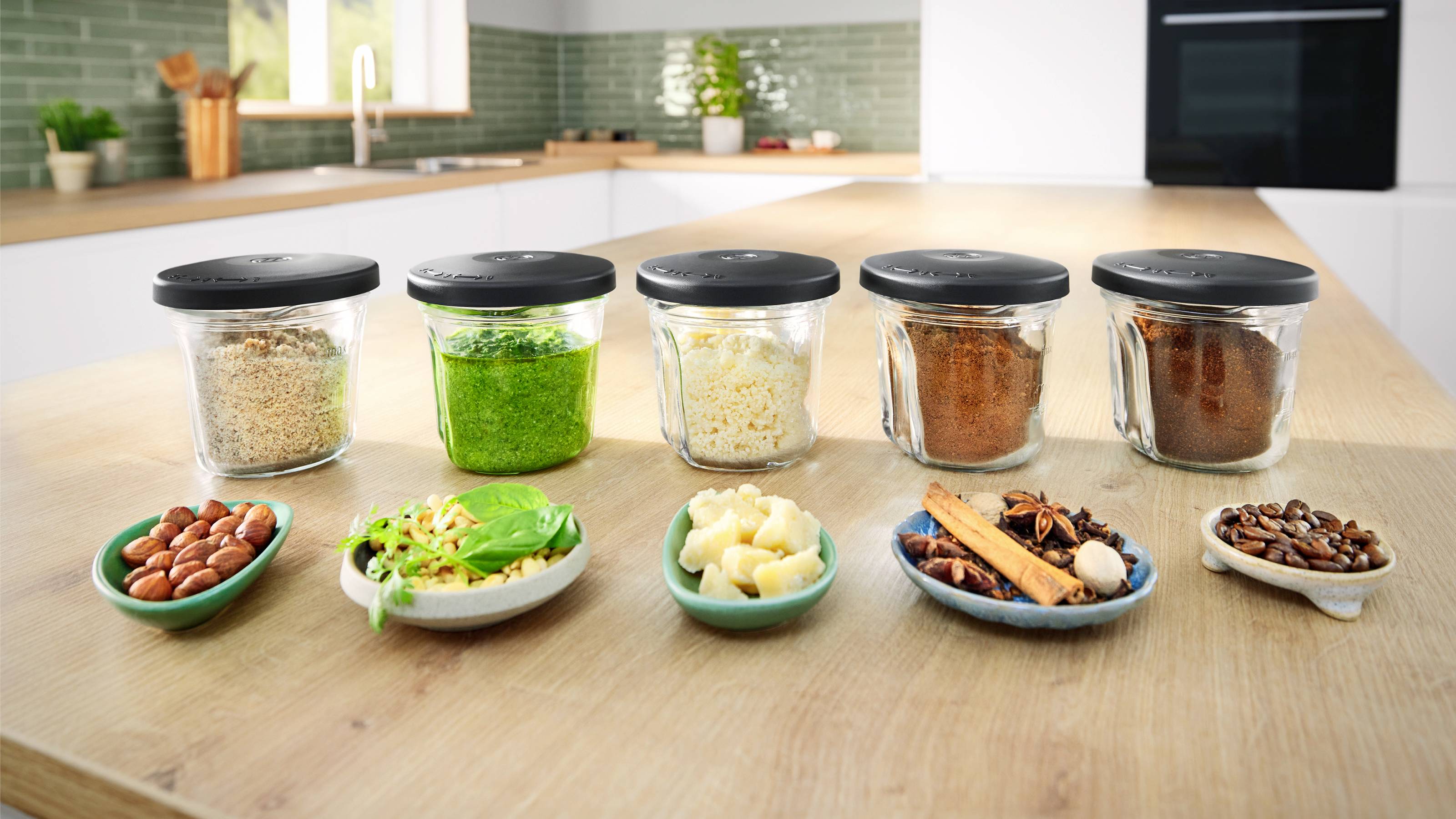 Fünf Gläser mit schwarzen Deckeln auf einer Küchenarbeitsplatte, gefüllt mit Reis, grünen Kräutern, Pasta, roten Gewürzen und Kakao, mit kleinen Schalen mit Zutaten davor.